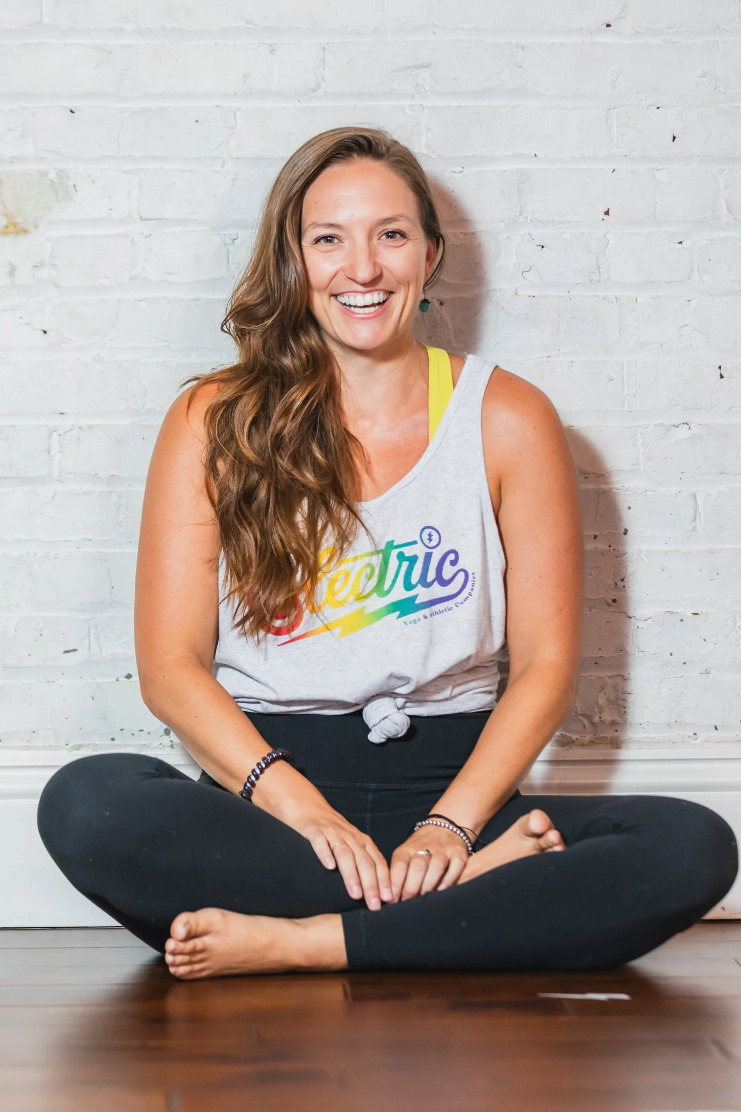 A smiling woman with long brown hair, sitting cross-legged on a wooden floor against a white brick wall. She is wearing a sleeveless white tank top with colorful text and black pants, and has a black beaded bracelet and earrings. She is in a yoga or meditation pose, with her hands resting on her knees and looking directly at the camera.
