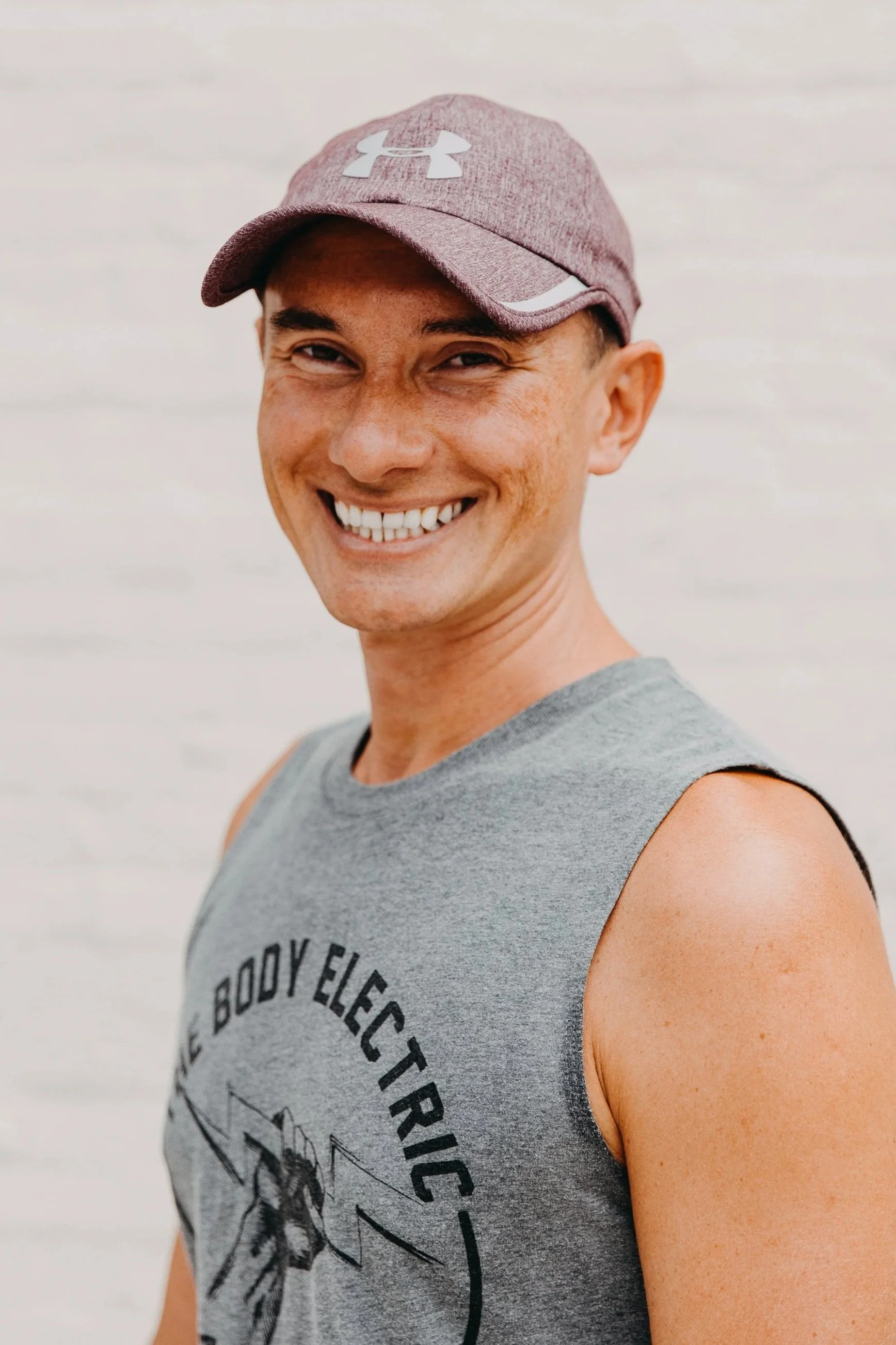 A smiling person wearing a maroon Under Armour cap and a gray sleeveless shirt with black text and graphics, standing against a light gray background.