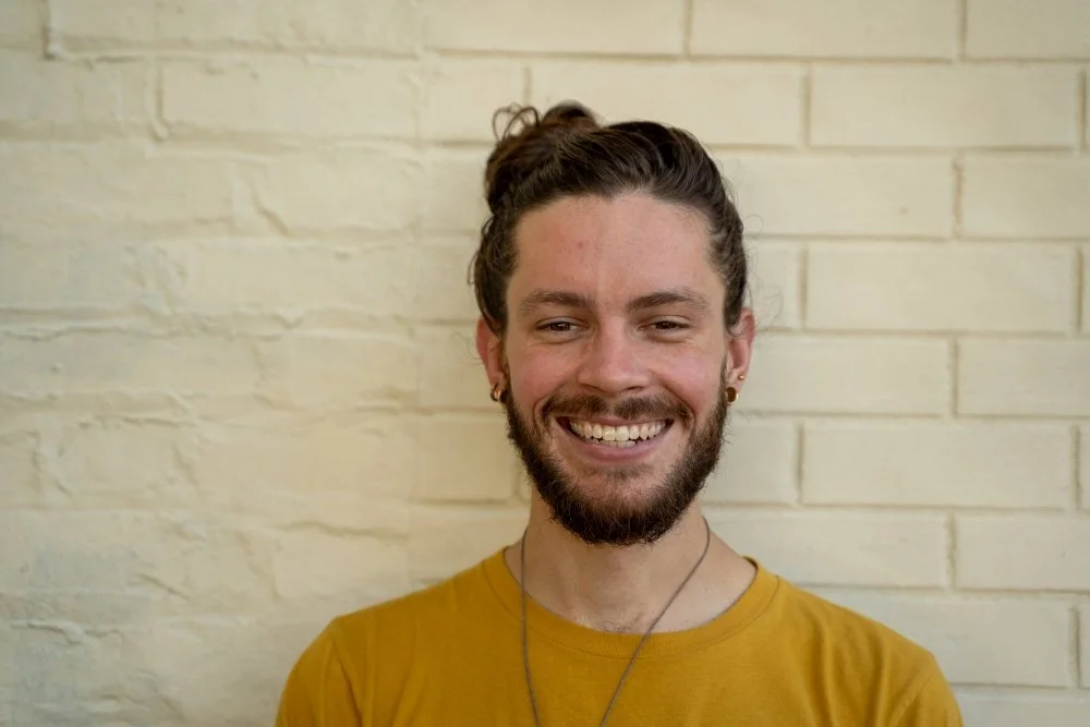 A young man with a beard smiling, wearing a yellow shirt and earrings, standing in front of a cream-colored brick wall.