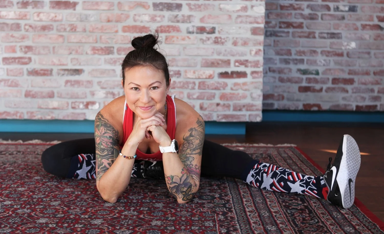 A woman with tattoos and dark hair tied in a bun shows a smile while doing a split on a patterned rug inside a room with a brick wall background.