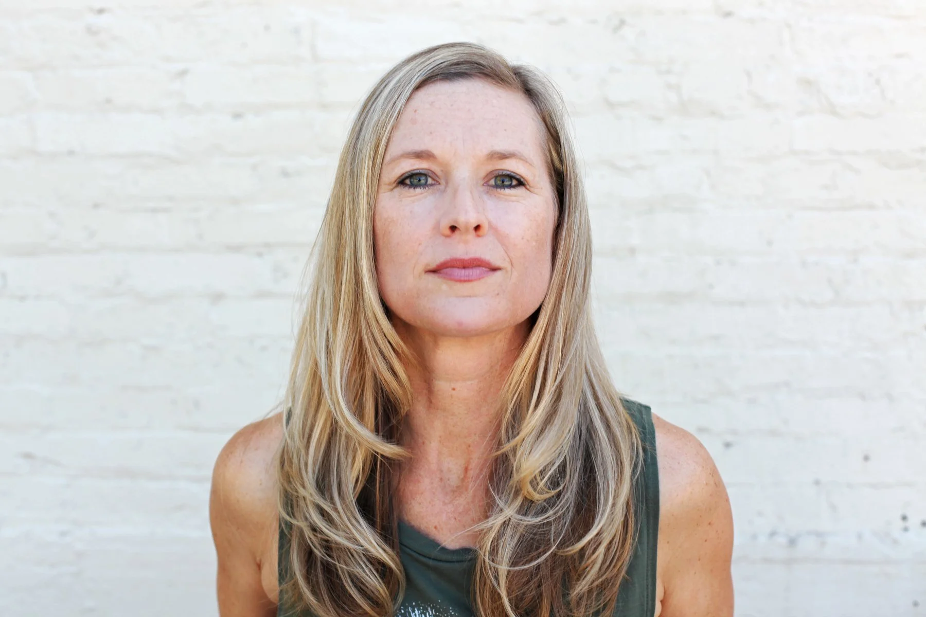 A woman with long blonde hair and blue eyes looking directly at the camera, standing in front of a white brick wall.
