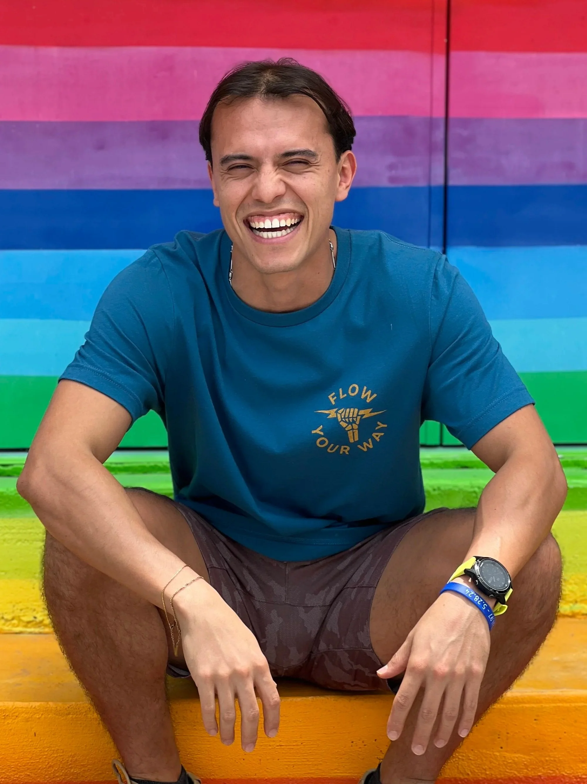 A smiling young man sitting on a rainbow-colored rainbow with rainbow stripes in the background. He is wearing a blue t-shirt, shorts, and a watch, with rainbow wristbands. The scene is vibrant and cheerful.