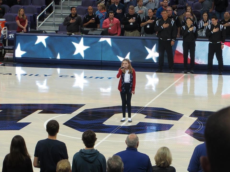 Young girl singing into a microphone on a basketball court during a national anthem performance, with spectators and team members standing with right hands over hearts in the background.