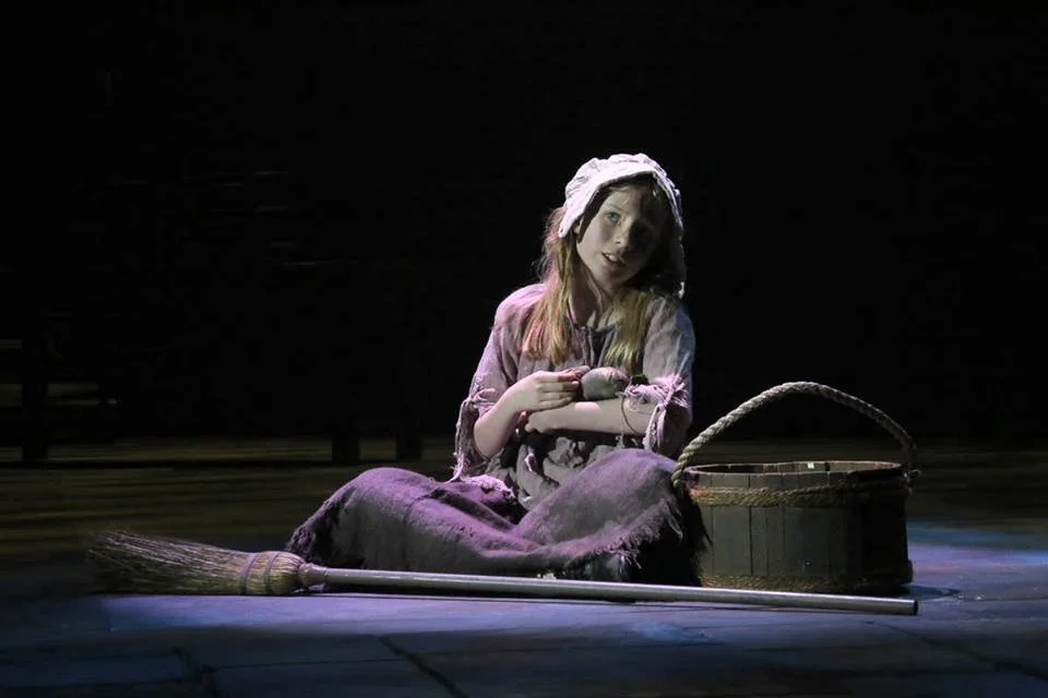 Young girl sitting on the floor with a broom, a basket, and a stuffed toy, wearing old-fashioned clothes and a bonnet, in a dark setting.