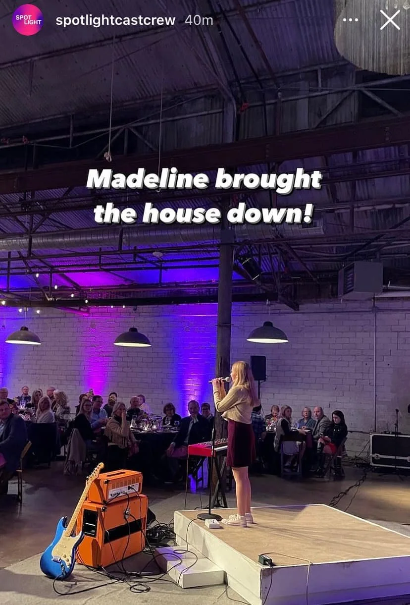A woman with long blonde hair wearing a beige jacket and dark skirt stands on a small stage, singing into a microphone. There are musical equipment and guitars on the stage. Behind her, an audience of people are seated at tables in a venue with white brick walls and industrial lighting. The photo appears to be taken during an event or performance.