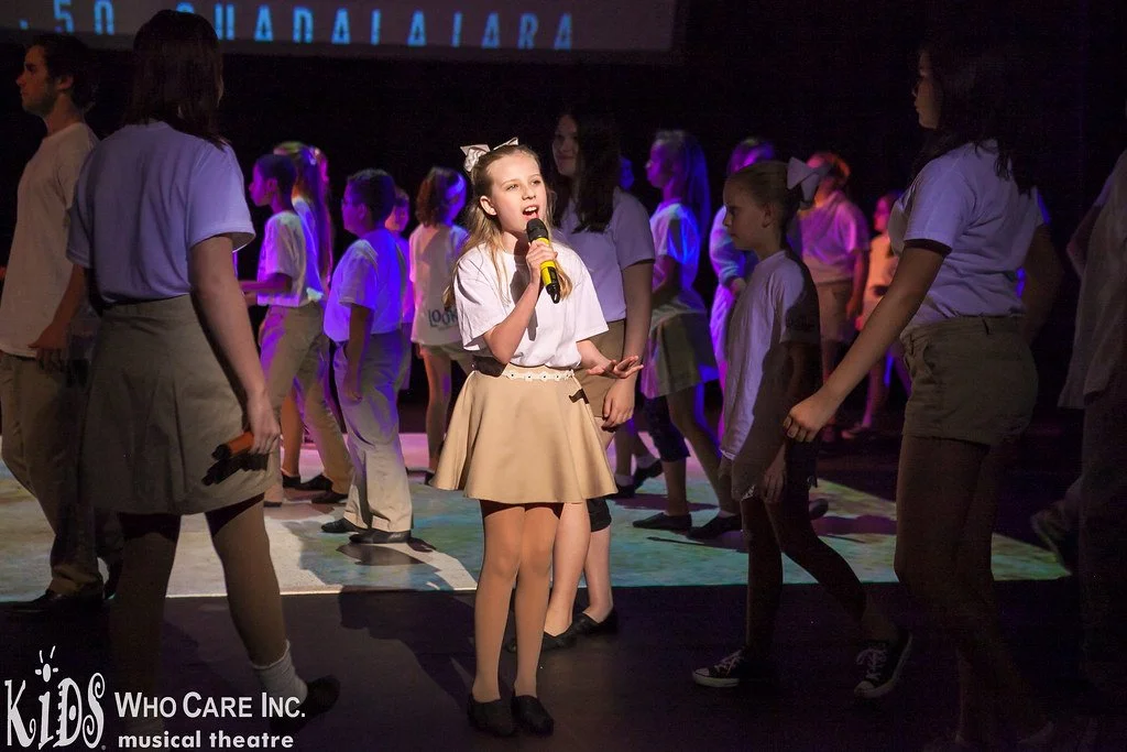 Young girl singing into a microphone on stage during a children's musical theater performance, with other children standing around her in costumes.