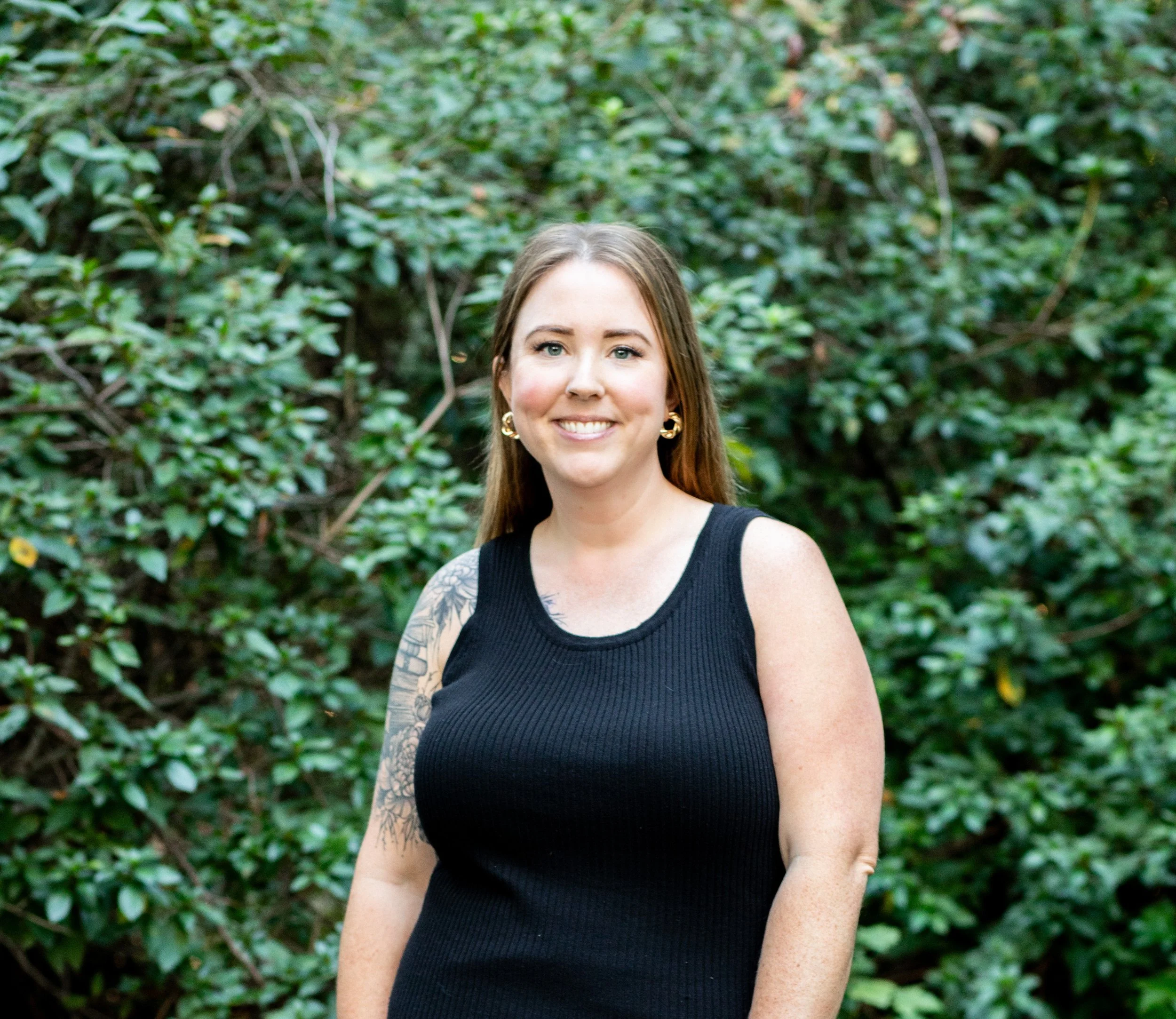 A smiling woman with long brown hair, gold hoop earrings, and a tattoo on her left shoulder, standing outdoors with green foliage in the background, wearing a sleeveless black top.