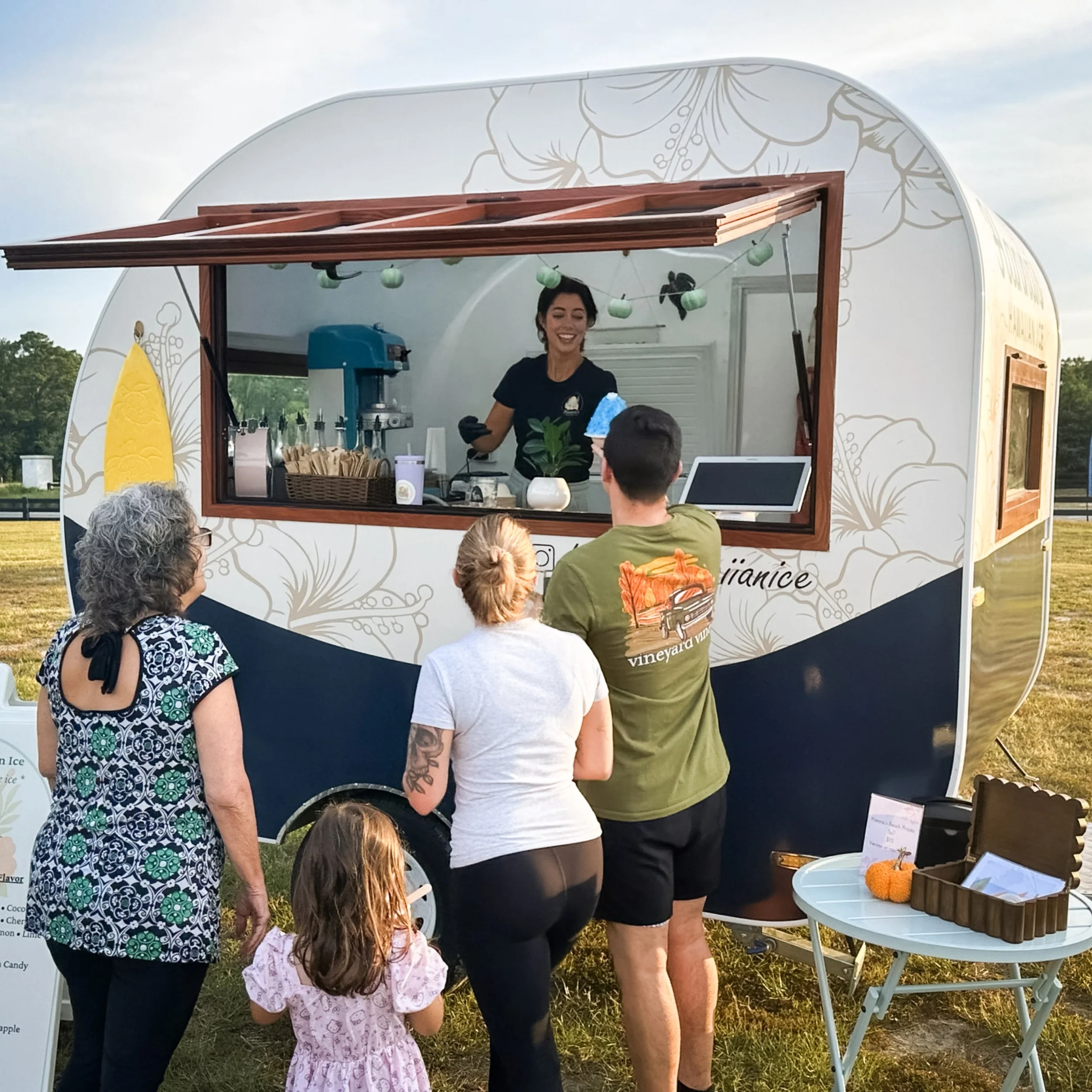 People ordering ice cream at a mobile ice cream truck during daytime with a smiling vendor inside making treats.