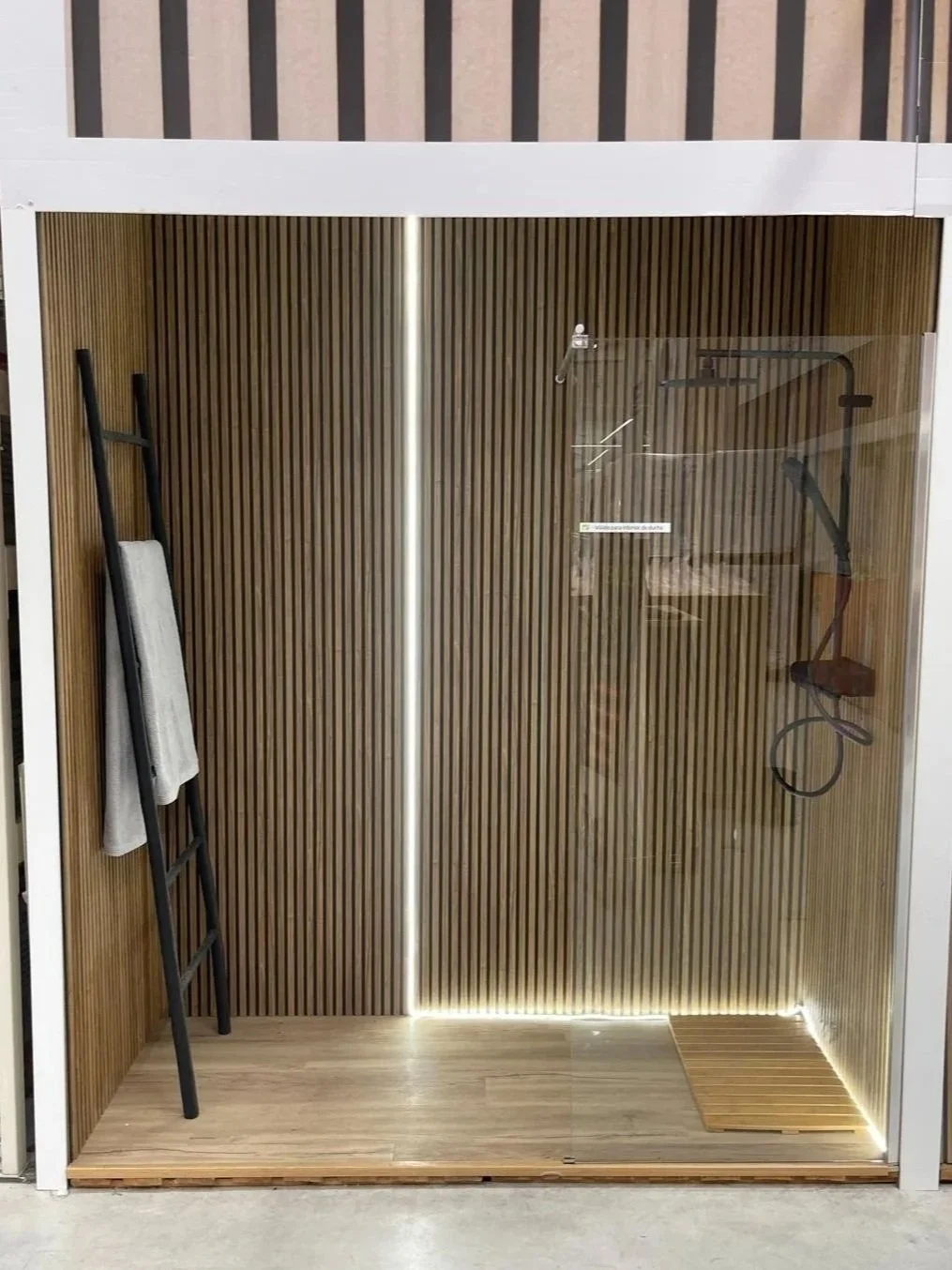 An outdoor shower enclosure with wood-paneled walls, a glass door, a towel hanging on a black ladder, and a wooden slatted floor mat.