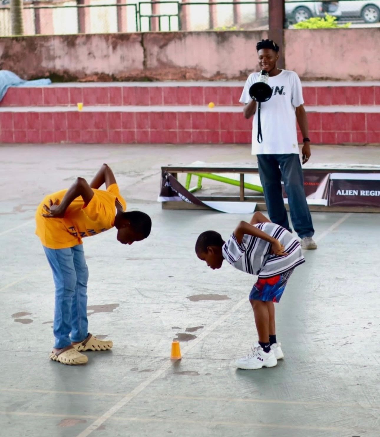Two boys bend over to look down, observing something on the ground, while a third boy stands nearby with a megaphone. A person in the background holds a camera, and there is a table and banners behind them. They are on an outdoor paved area.