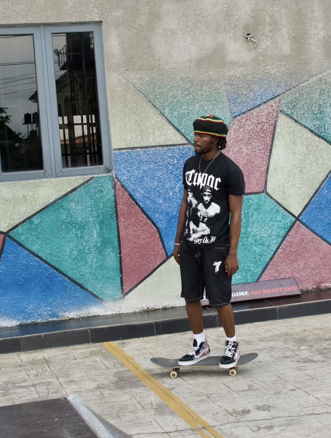 A young man skateboarding in front of a colorful geometric painted wall, wearing a black graphic T-shirt, black shorts, and a multicolored beanie.