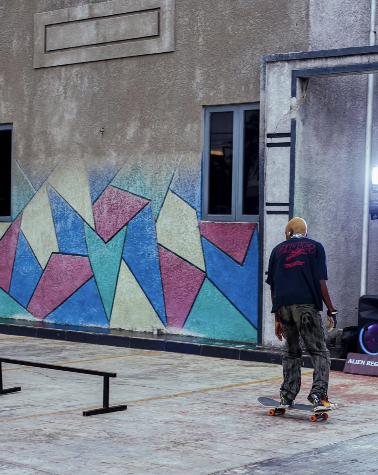 A person skateboarding on a concrete surface next to a colorful geometric mural on a building wall, with a window and an open doorway visible.
