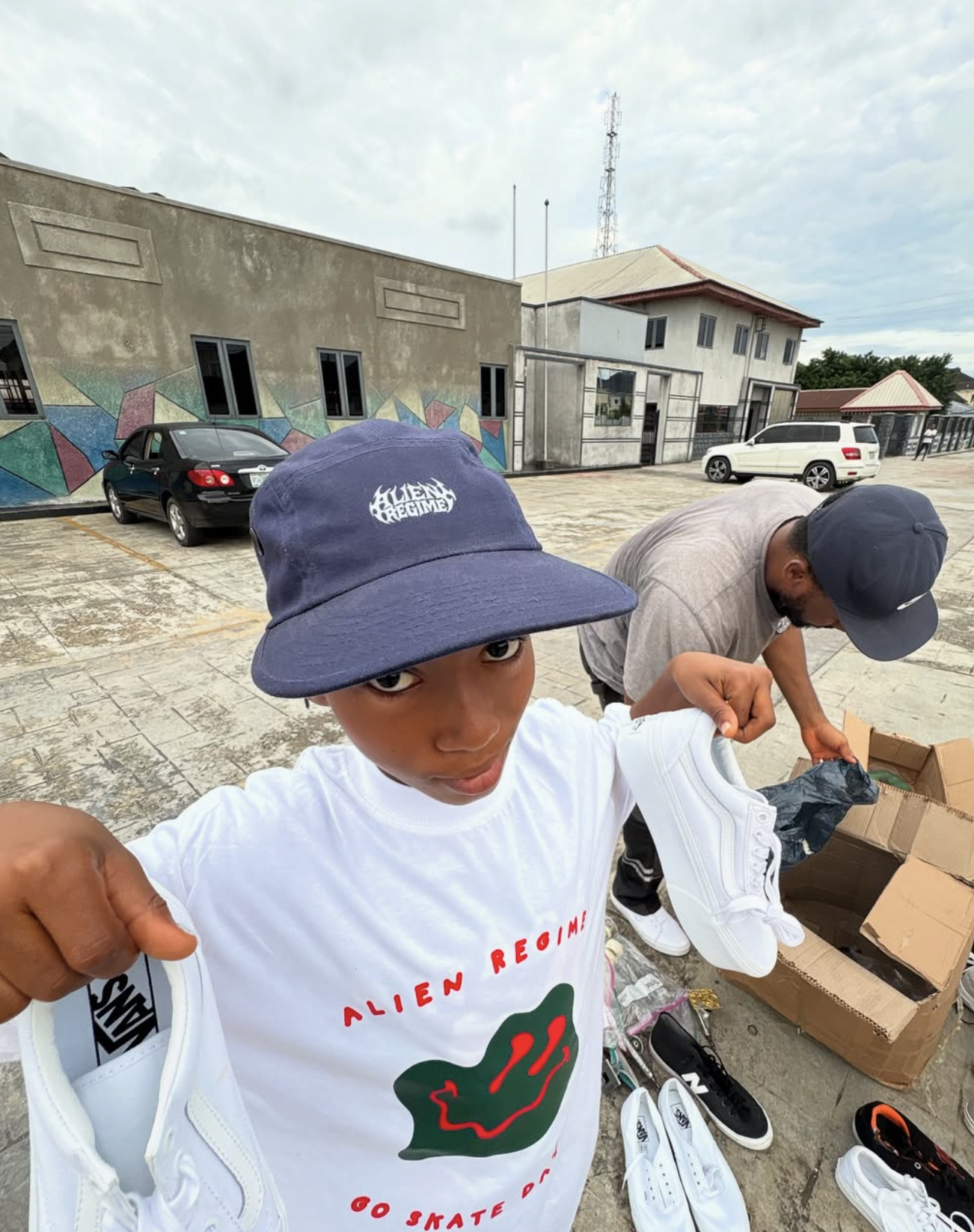 Young boy wearing a navy bucket hat and white t-shirt holding a pair of white sneakers, with a man in the background sorting shoes in an outdoor parking lot.