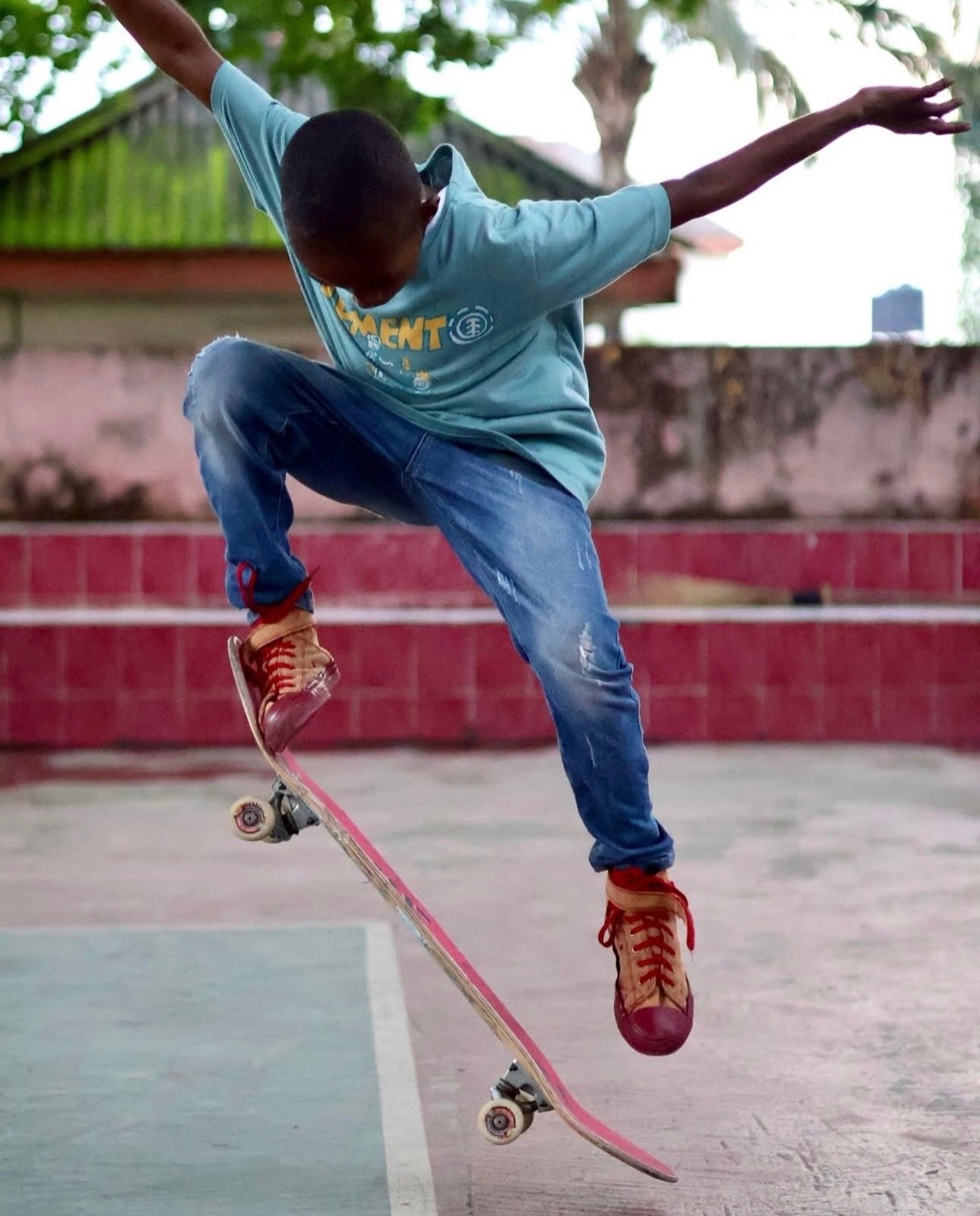 A person skateboarding outdoors, mid-air, wearing a light blue T-shirt, ripped blue jeans, and red high-top sneakers.