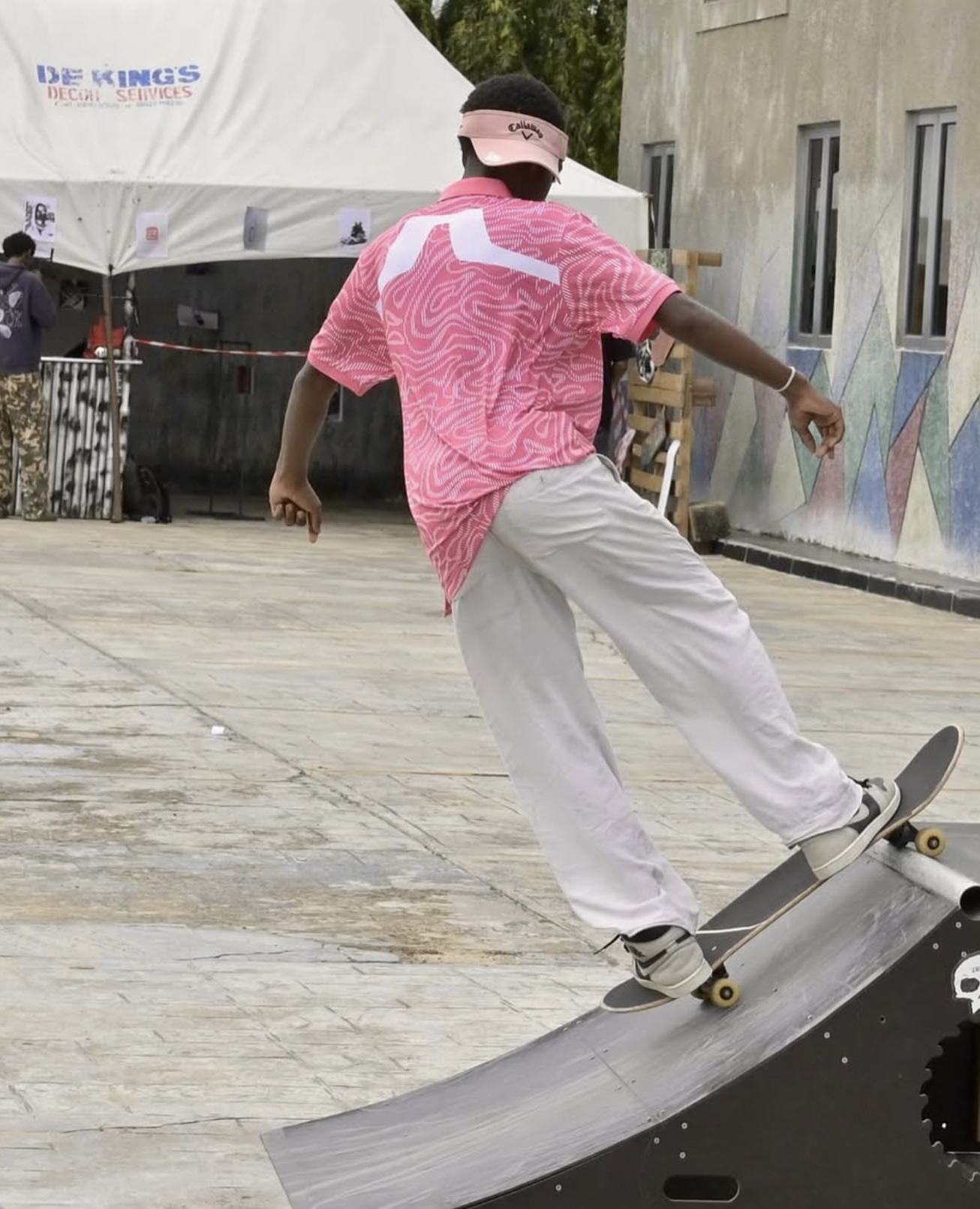 A young person wearing a pink cap, pink shirt, and white pants skateboarding on a ramp outdoors.