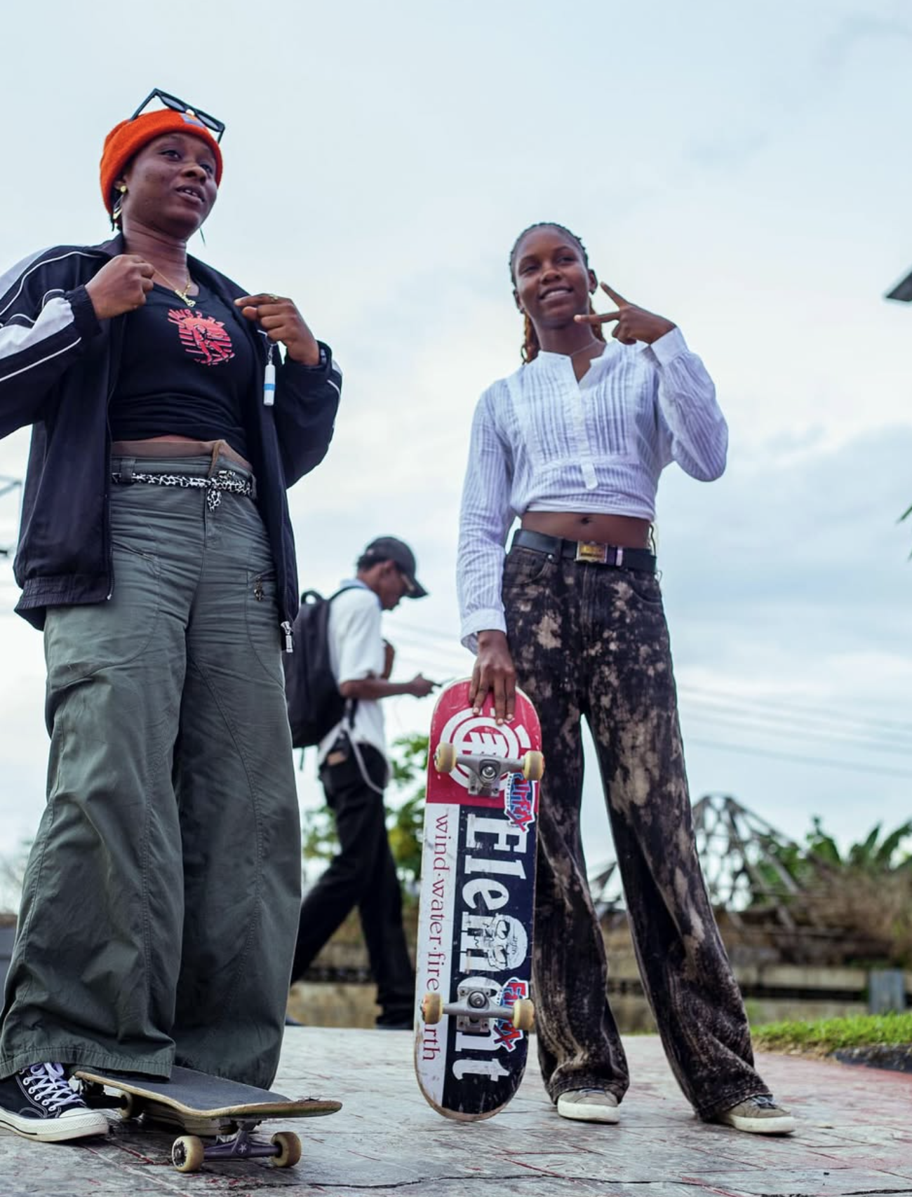 Two young women with skateboards outdoors, one holding a skateboard and the other making a peace sign, with a man in the background looking at his phone.