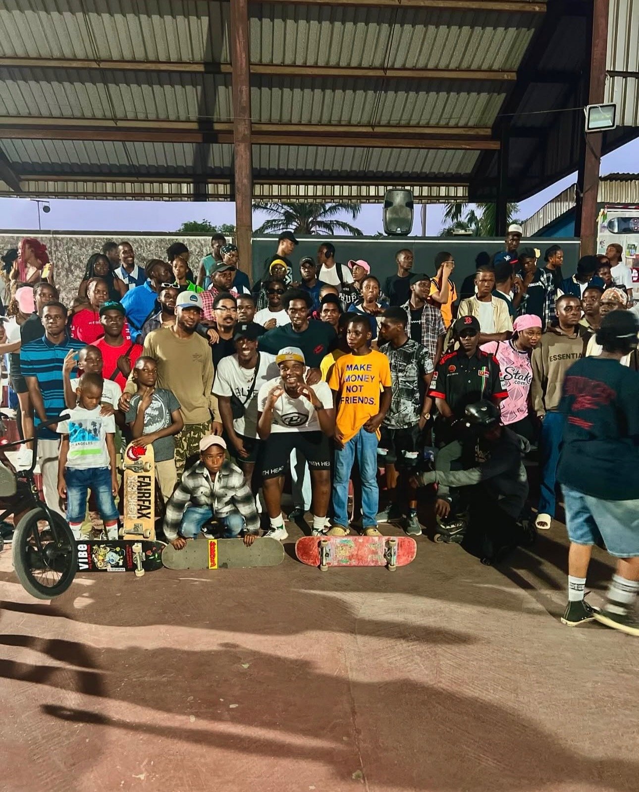 A large group of people, including children and adults, gathered under a covered outdoor area. Some are holding skateboards, and there is a bicycle visible on the left side. The setting appears to be a skate park or similar recreational area.