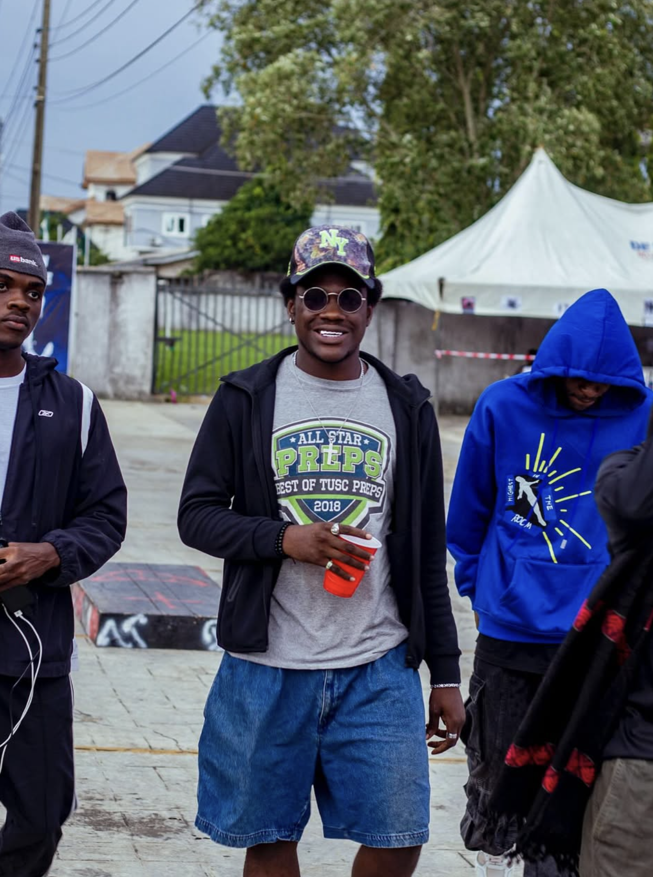 A group of young men are standing outdoors on a paved area. The man in the center is smiling, wearing round sunglasses, a baseball cap with a New York logo, a gray t-shirt, a black jacket, and denim shorts. He is holding a red plastic cup. There is a