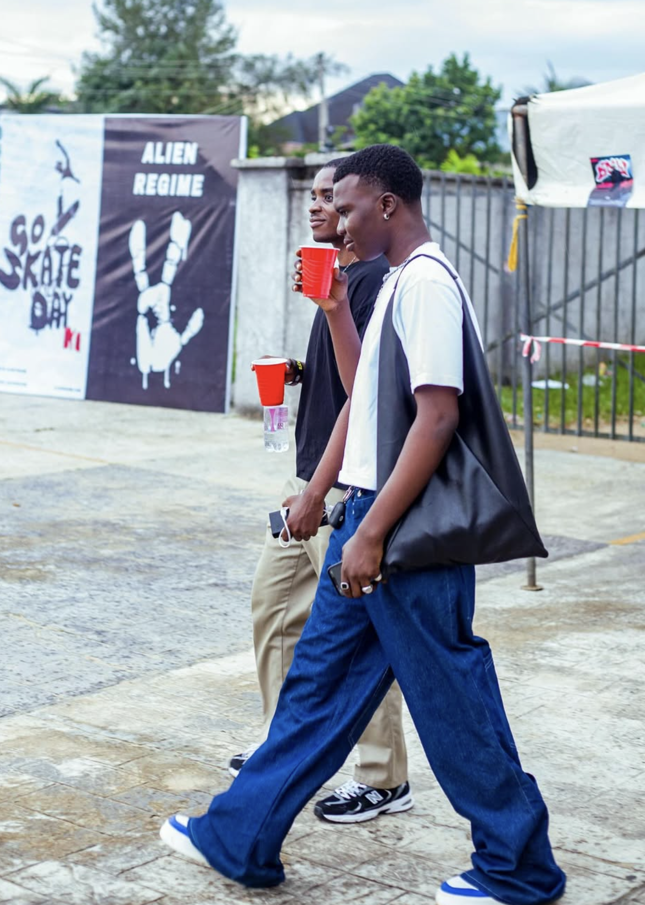 Two young men walking outdoors near a fence and a banner with the text 'ALIEN REGIME' and 'GO SKATE DAY'. One is holding a red cup and a phone, and the other is holding a red cup and a water bottle.