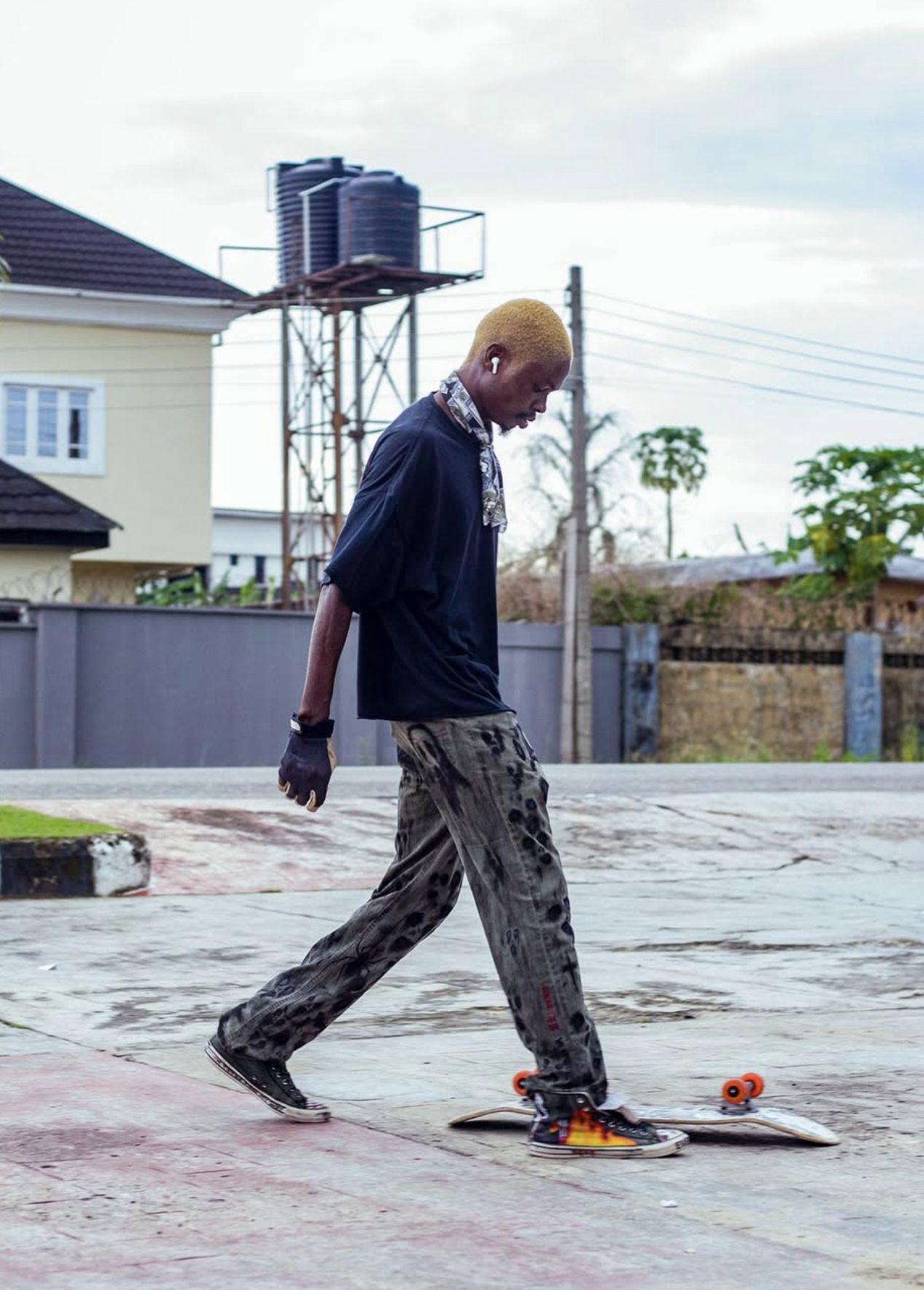Young man with blond hair walking on a skateboard on a concrete surface outdoors, wearing black t-shirt, patterned pants, sneakers, and wireless earbud.