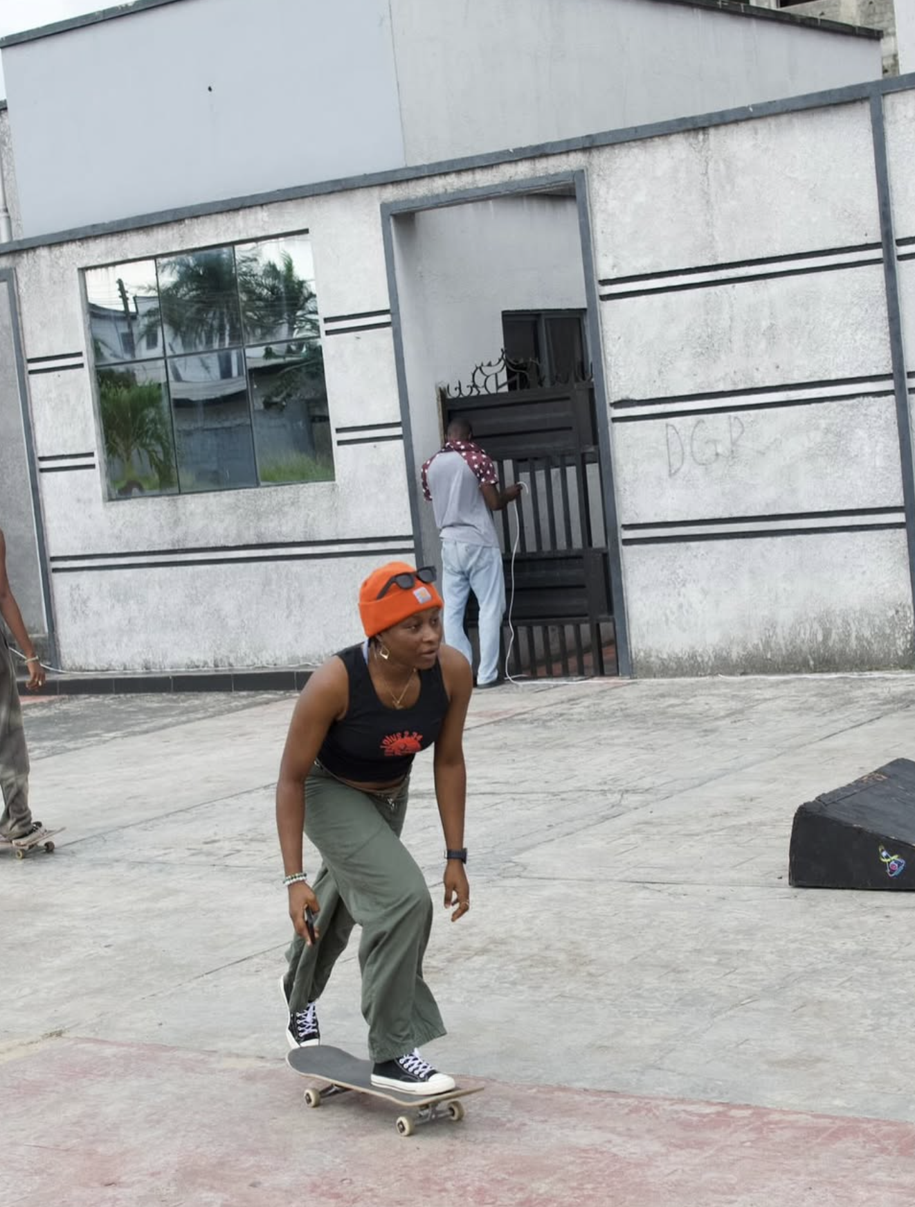 A woman wearing a black tank top, green pants, and a bright orange hat skateboards outdoors on a concrete surface. A man in a gray shirt with red and white sleeves is seen in the background near a black gate, and another person is partially visible o
