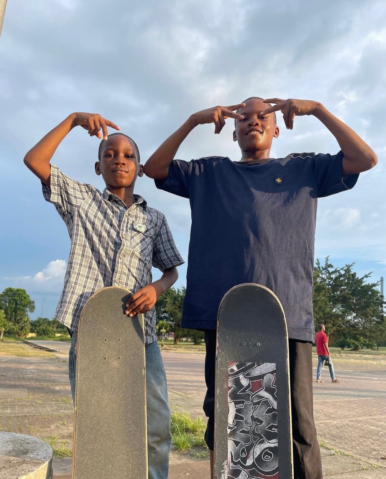 Two boys standing outdoors with skateboards, making peace signs with their hands, under a cloudy sky.