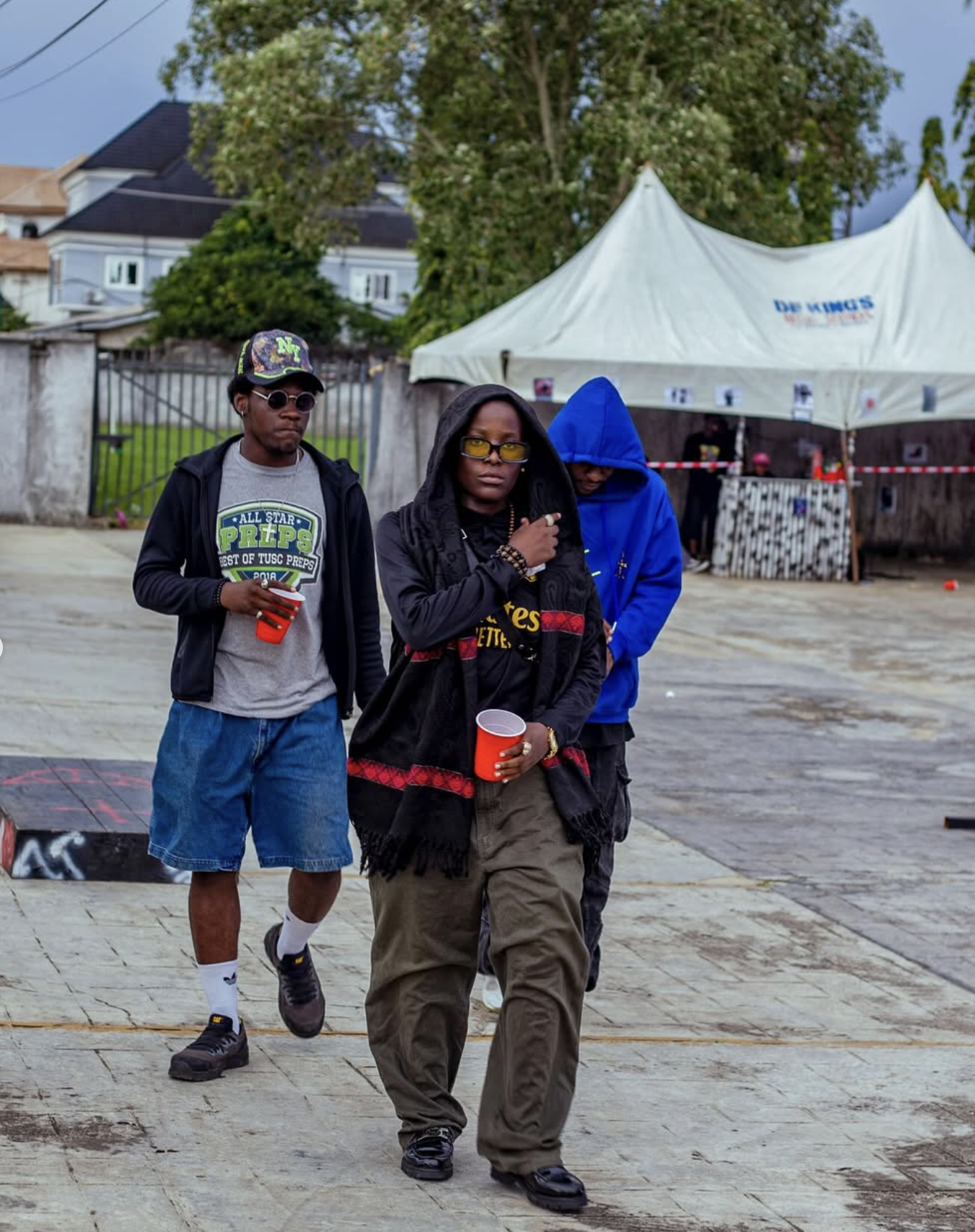Three young people standing outdoors, two men and one woman, in casual attire holding red cups at an event with a white tent in the background.