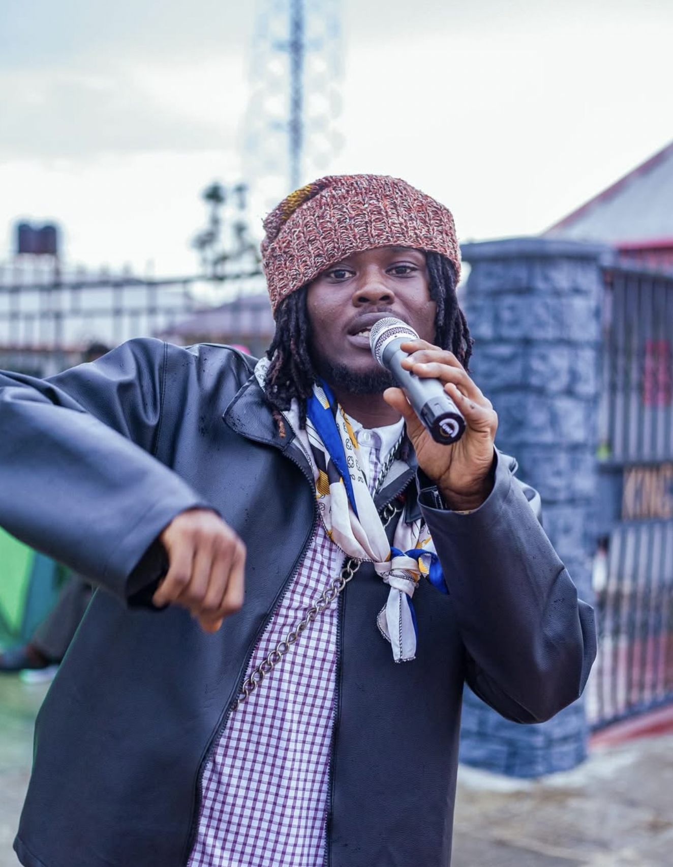 A man wearing a multicolored knit cap, black leather jacket, and patterned shirt, holding a microphone and pointing with his other hand, speaking outdoors.