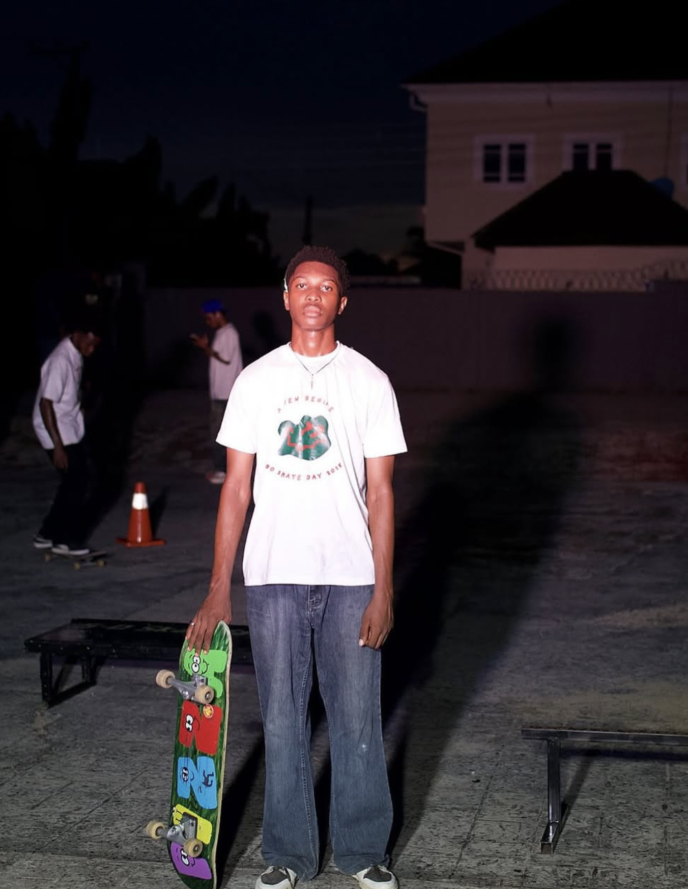 Young man standing outdoors at night holding a skateboard, wearing a white T-shirt and baggy jeans, with a dark sky and other people skateboarding in the background.