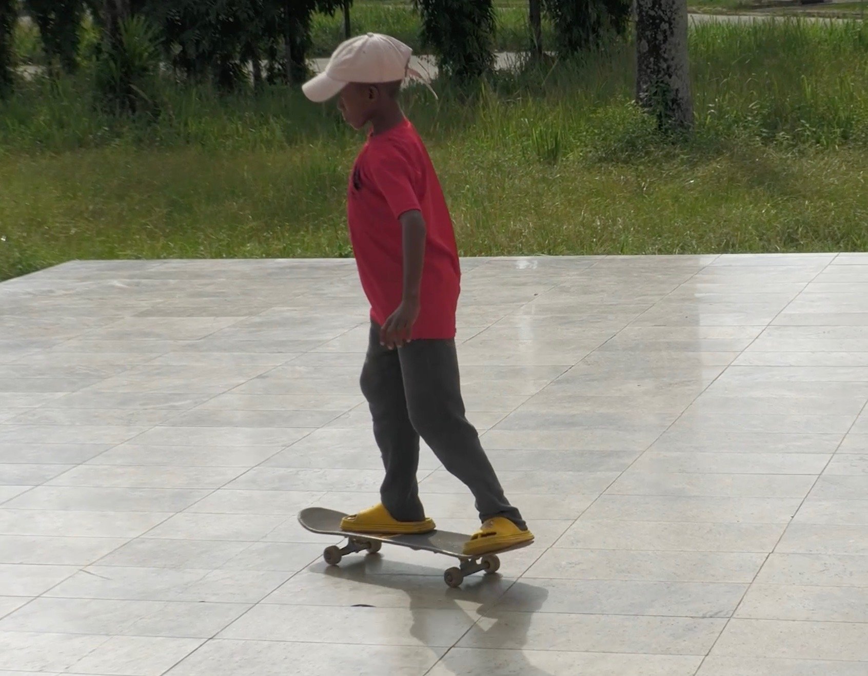 Child skateboarding outdoors on a tiled surface, wearing a pink shirt, gray pants, yellow slippers, and a white cap.
