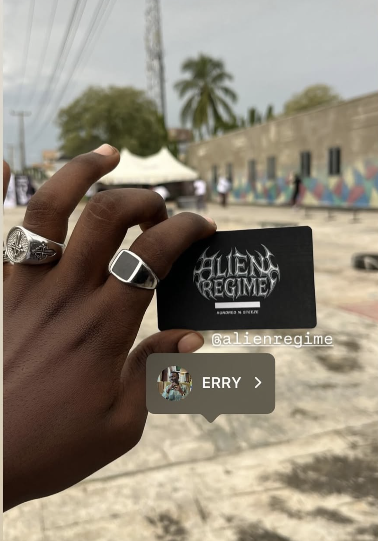 A person holding a black card with 'Alien Regime' written on it, with a background of an outdoor area with a colorful mural on a wall, palm trees, and a tent.