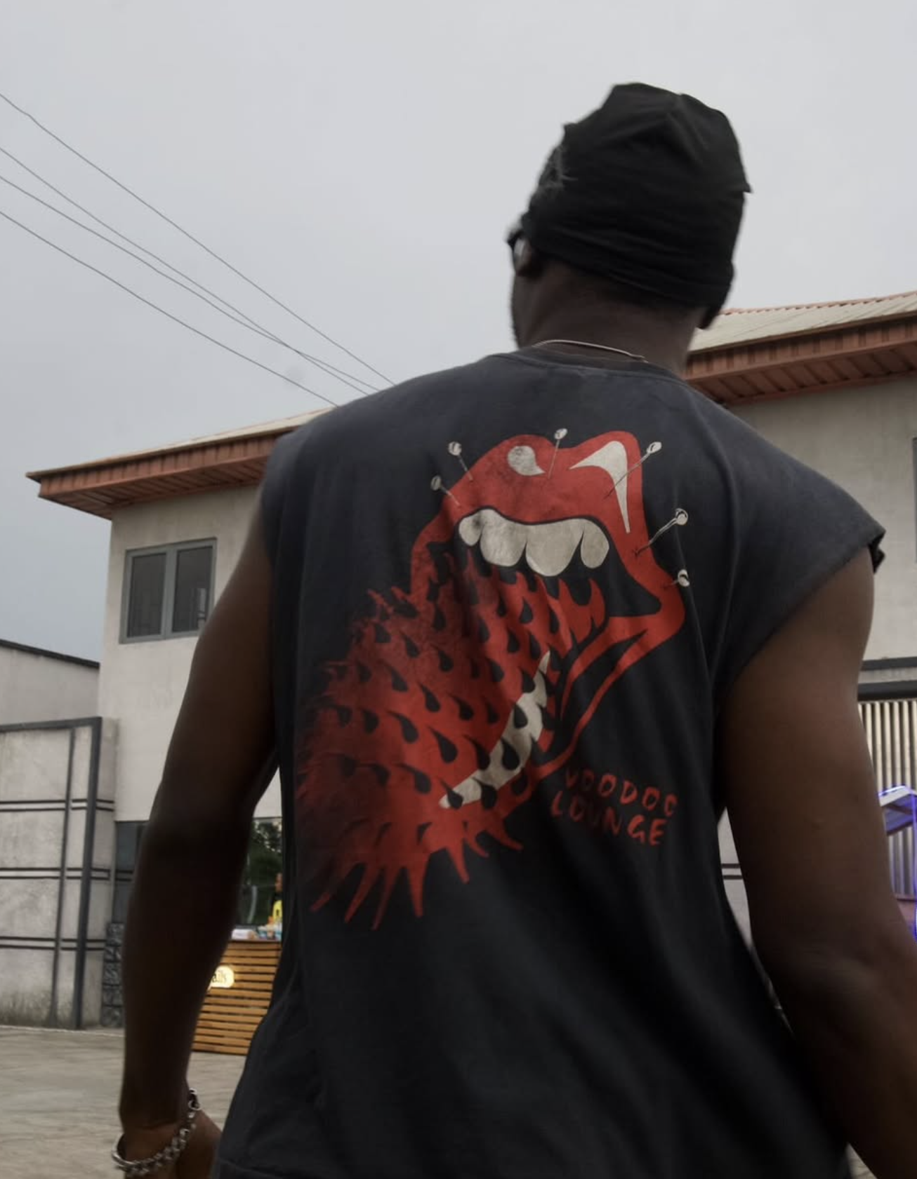 Back view of a person wearing a black sleeveless t-shirt with large red graphic of a mouth with sharp teeth, tongue, and surrounding pins, with the text 'Voodoo Lounge', outdoors in daytime.