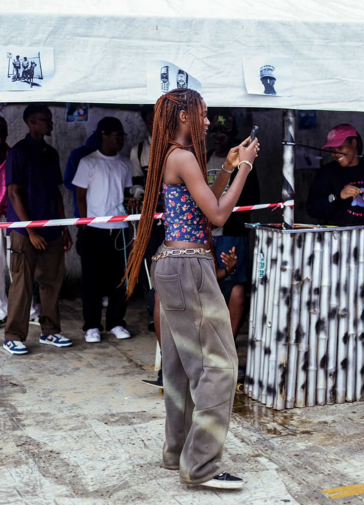 A young woman with long braided hair and casual clothing is taking a photo with her phone at an outdoor event. Several people are standing behind a barrier under a makeshift tent.