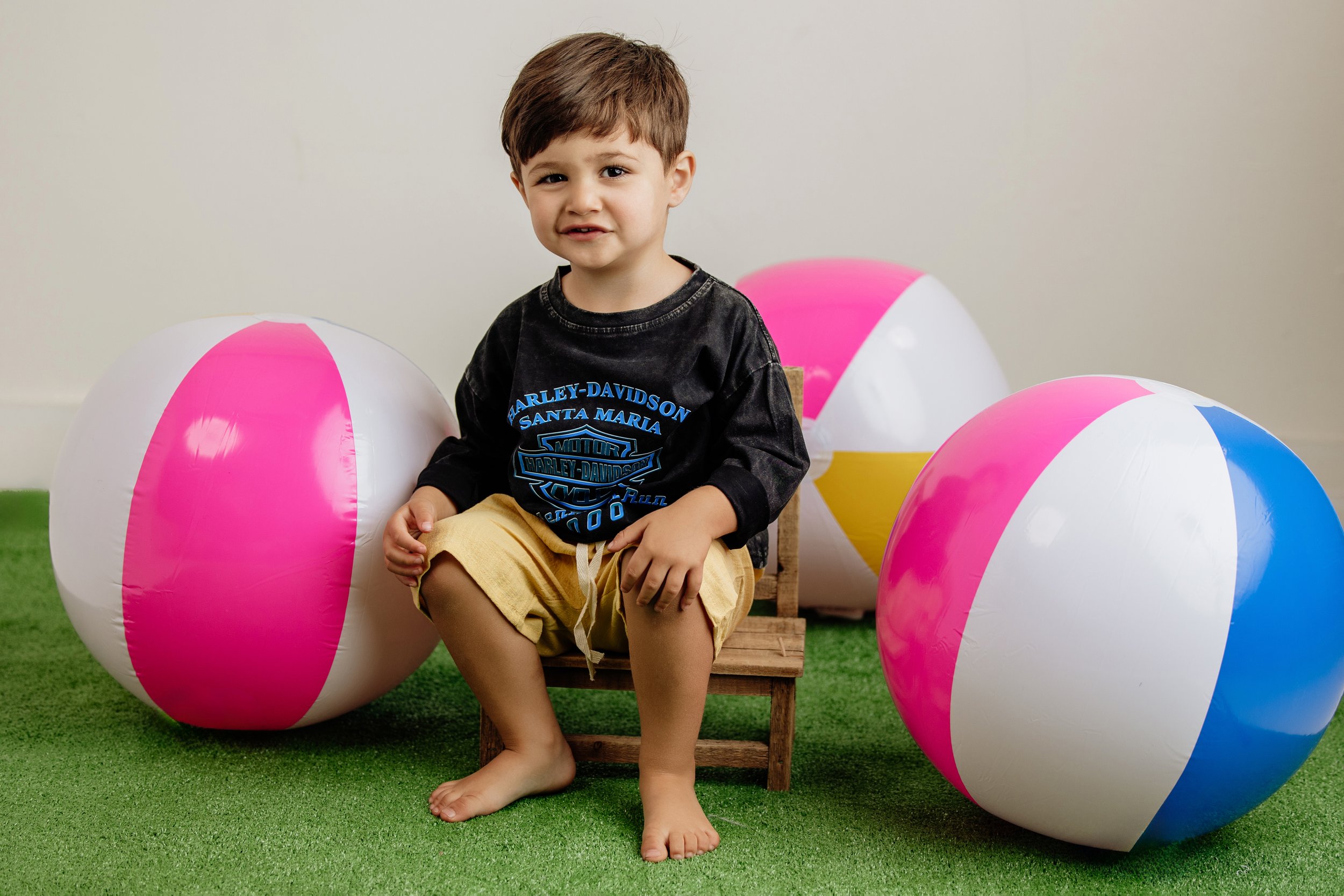Toddler boy with brown hair is wearing a Harley shirt and yellow shorts from North Georgia children's clothing store. He is sitting on a small bench in between colorful beachballs.