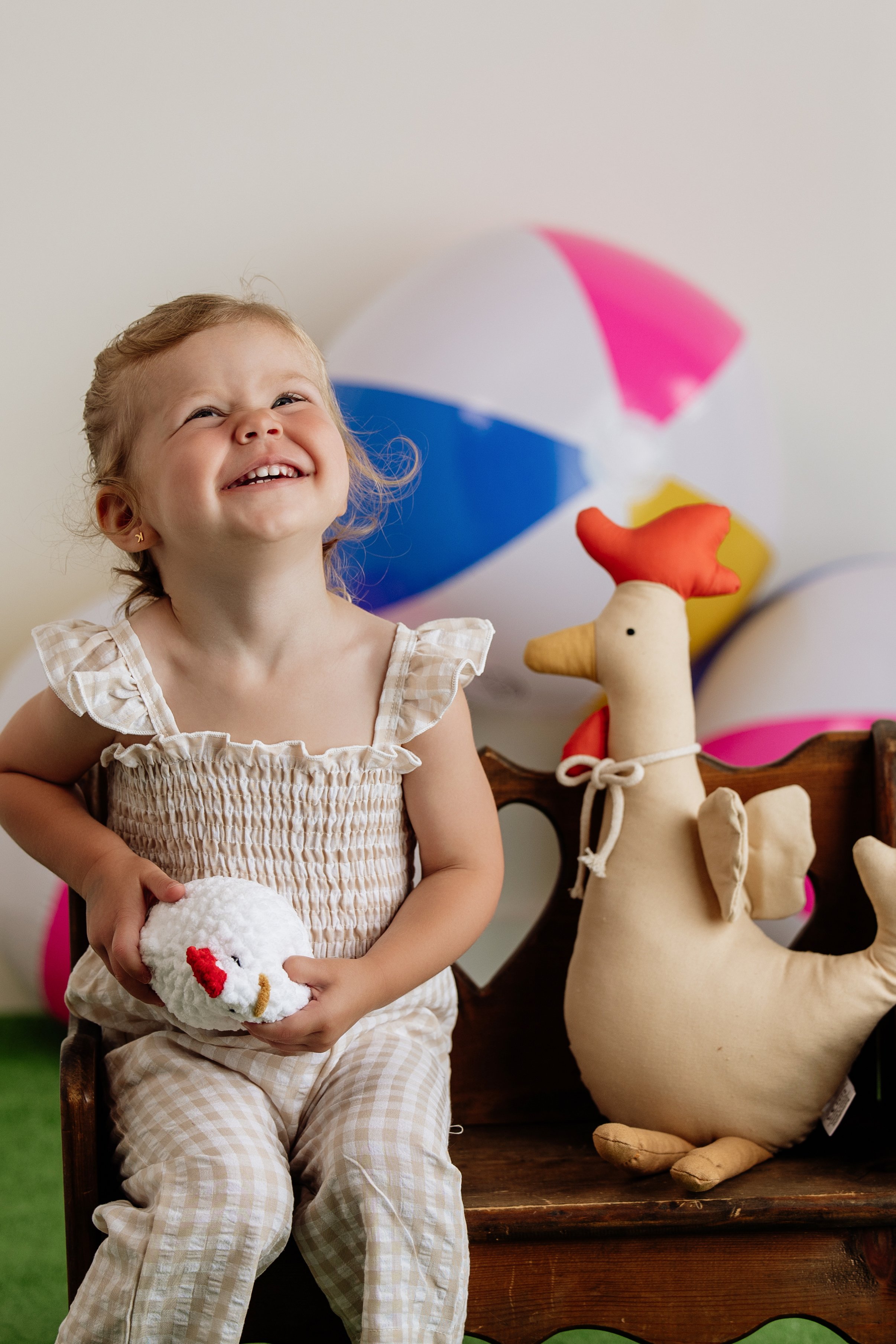 Little girl smiling on a wooden bench with a beach ball backdrop holding a plush chicken. She is wearing a gingham jumpsuit from local children's boutique on the historic square in Dahlonega, GA