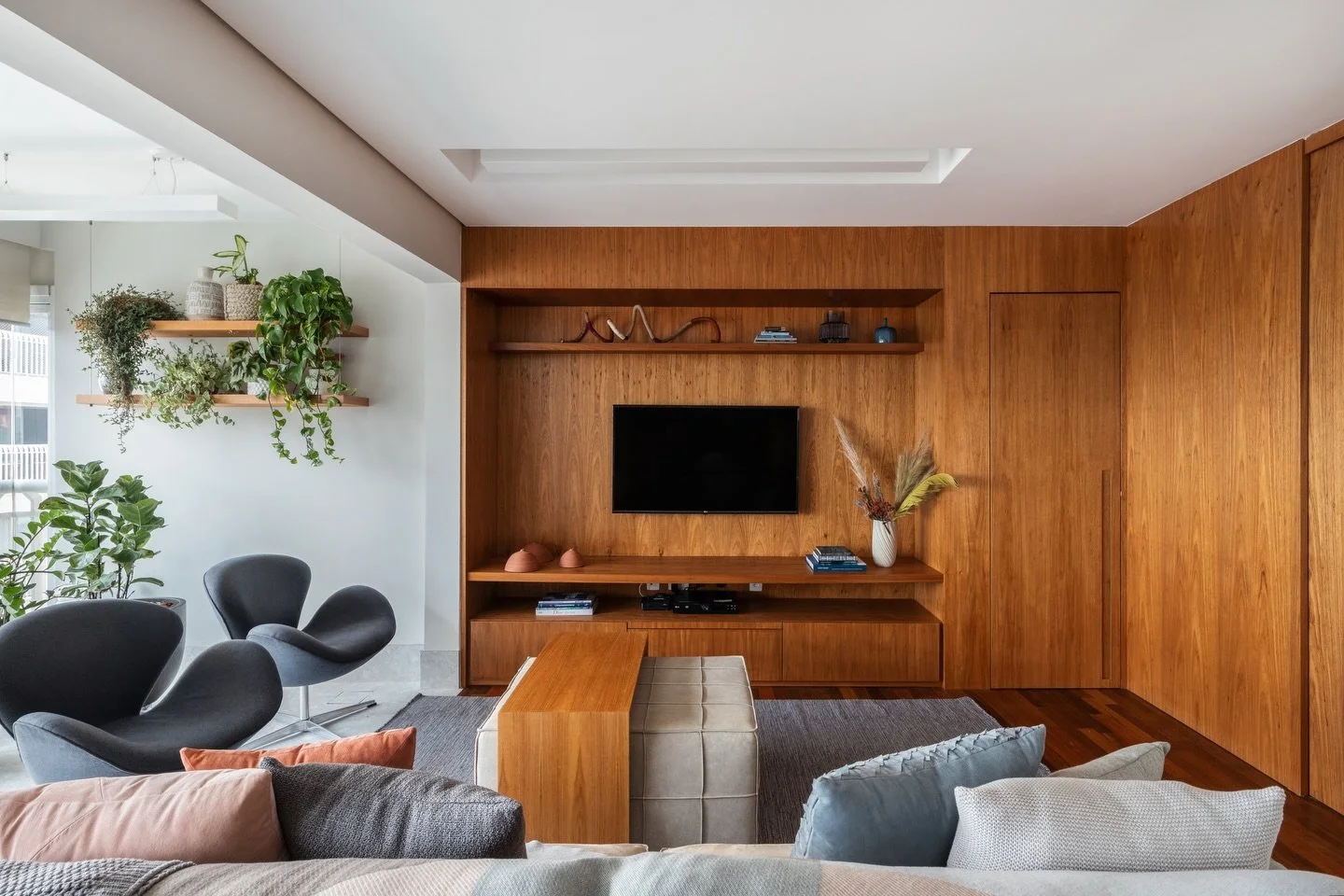 Nesse ambiente, a madeira aparece como elemento principal e organiza o espa&ccedil;o sem pesar. A estante integra TV, armazenamento e circula&ccedil;&atilde;o, enquanto a luz natural e o uso de plantas trazem equil&iacute;brio e conforto para o dia a