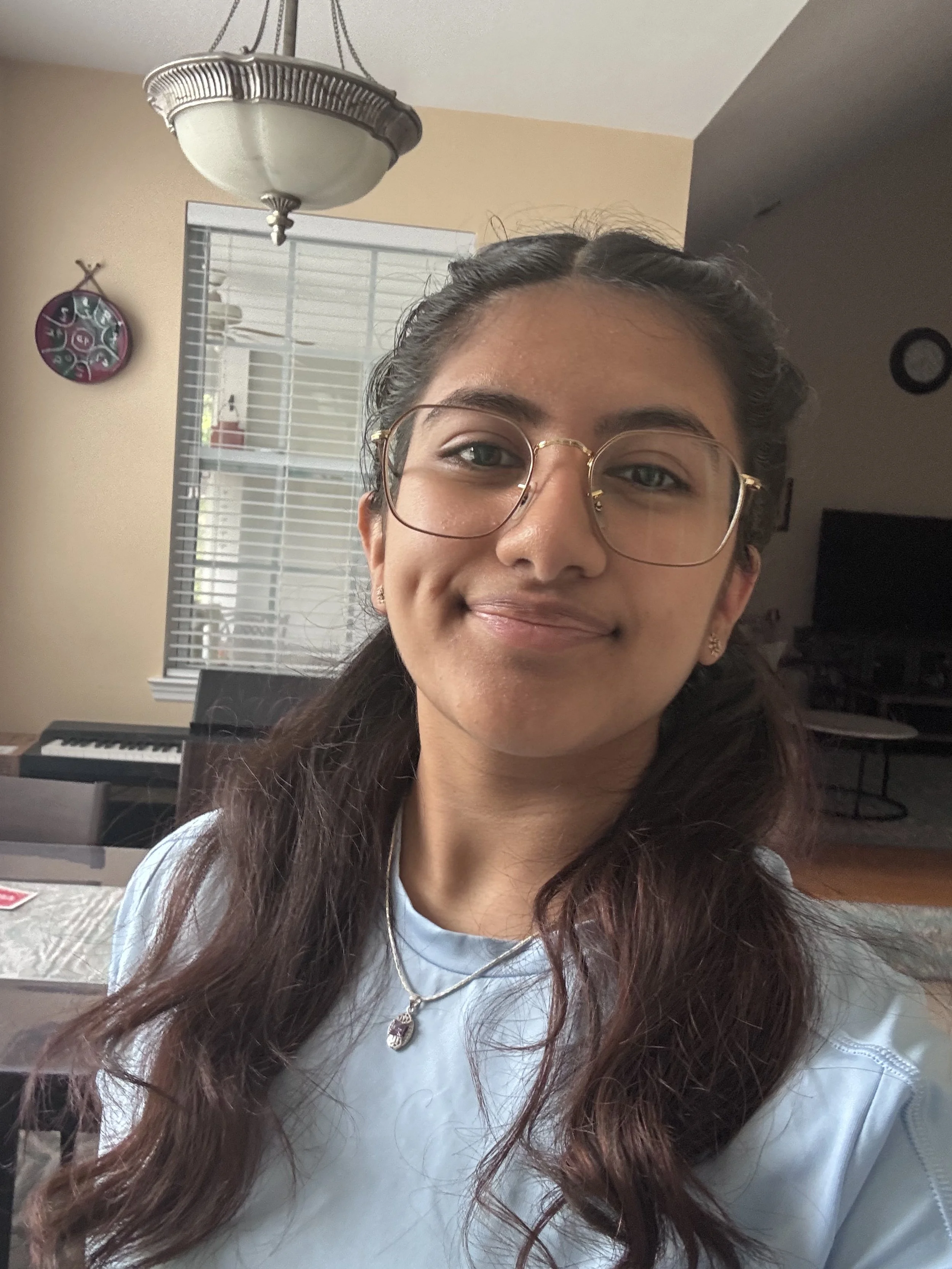 A young woman with glasses and long wavy hair smiling indoors, with a piano and window in the background.