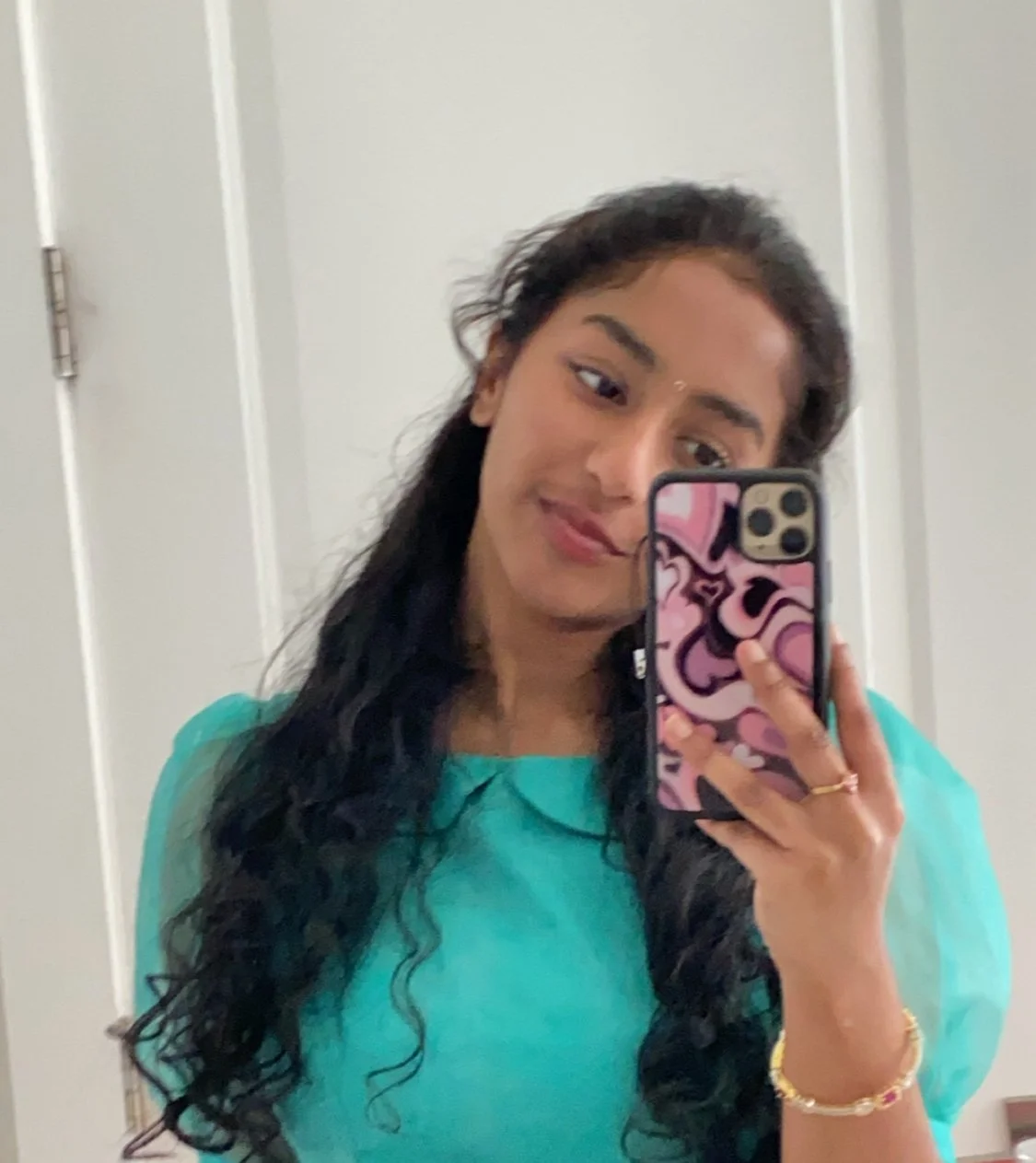 Young woman taking a mirror selfie with a smartphone, wearing a turquoise top and gold jewelry, with long wavy black hair, in a room with white walls and a door in the background.