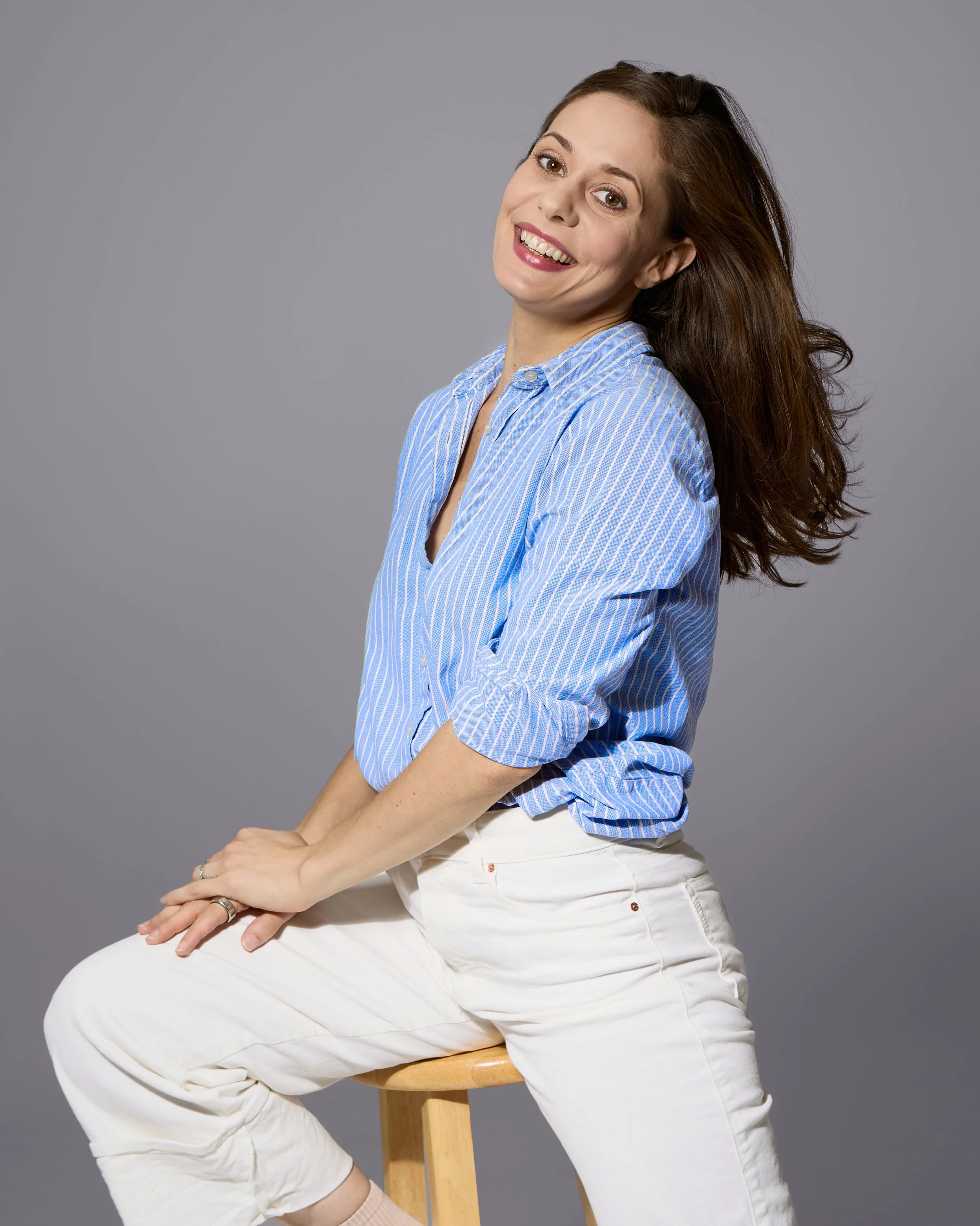 A woman with long brown hair and a bright smile, dressed in a light blue striped shirt and white pants, sitting on a wooden stool against a plain gray background.