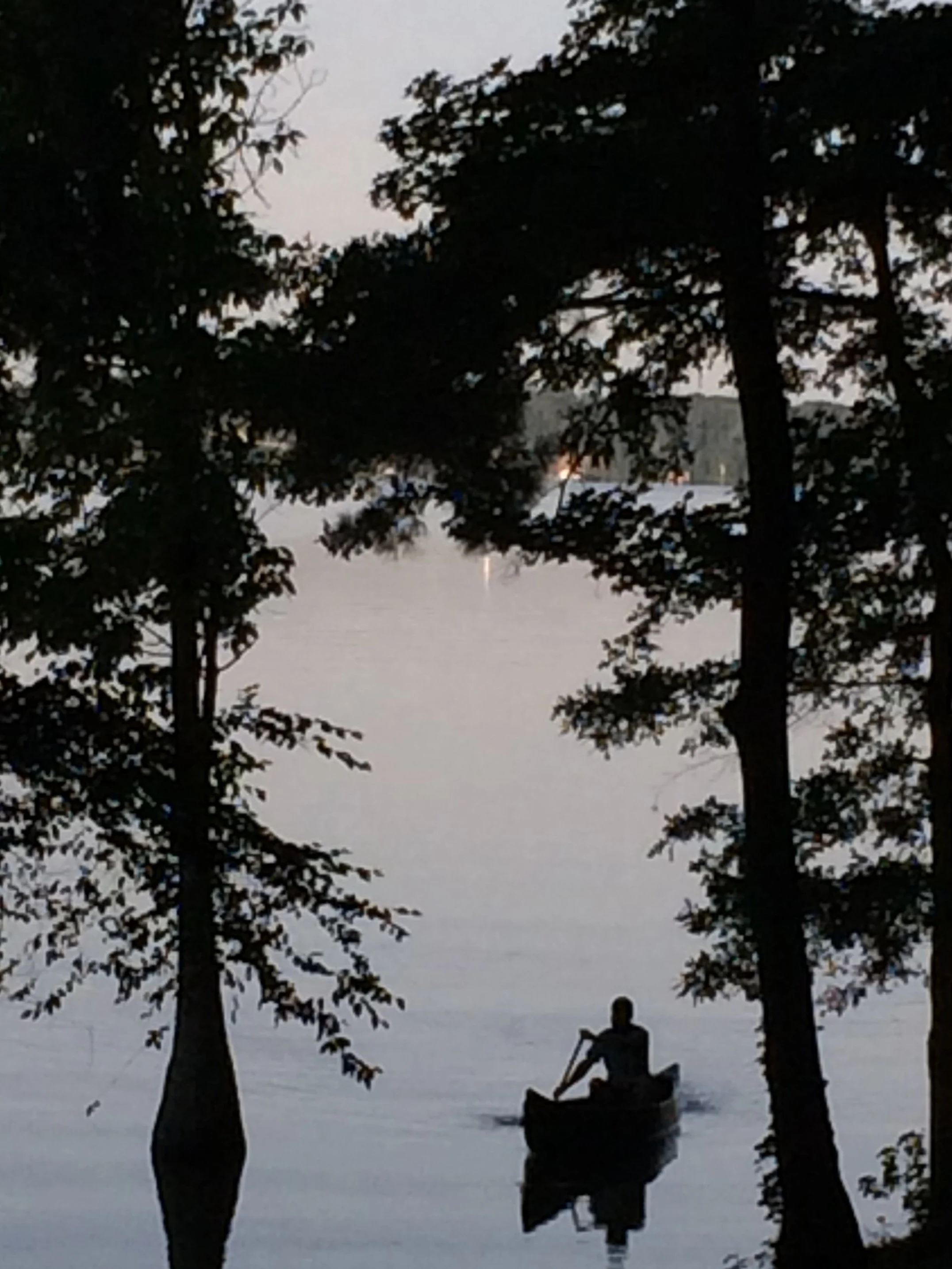 A person kayaking on a calm body of water surrounded by trees, with their silhouette visible against the water at dusk or dawn.