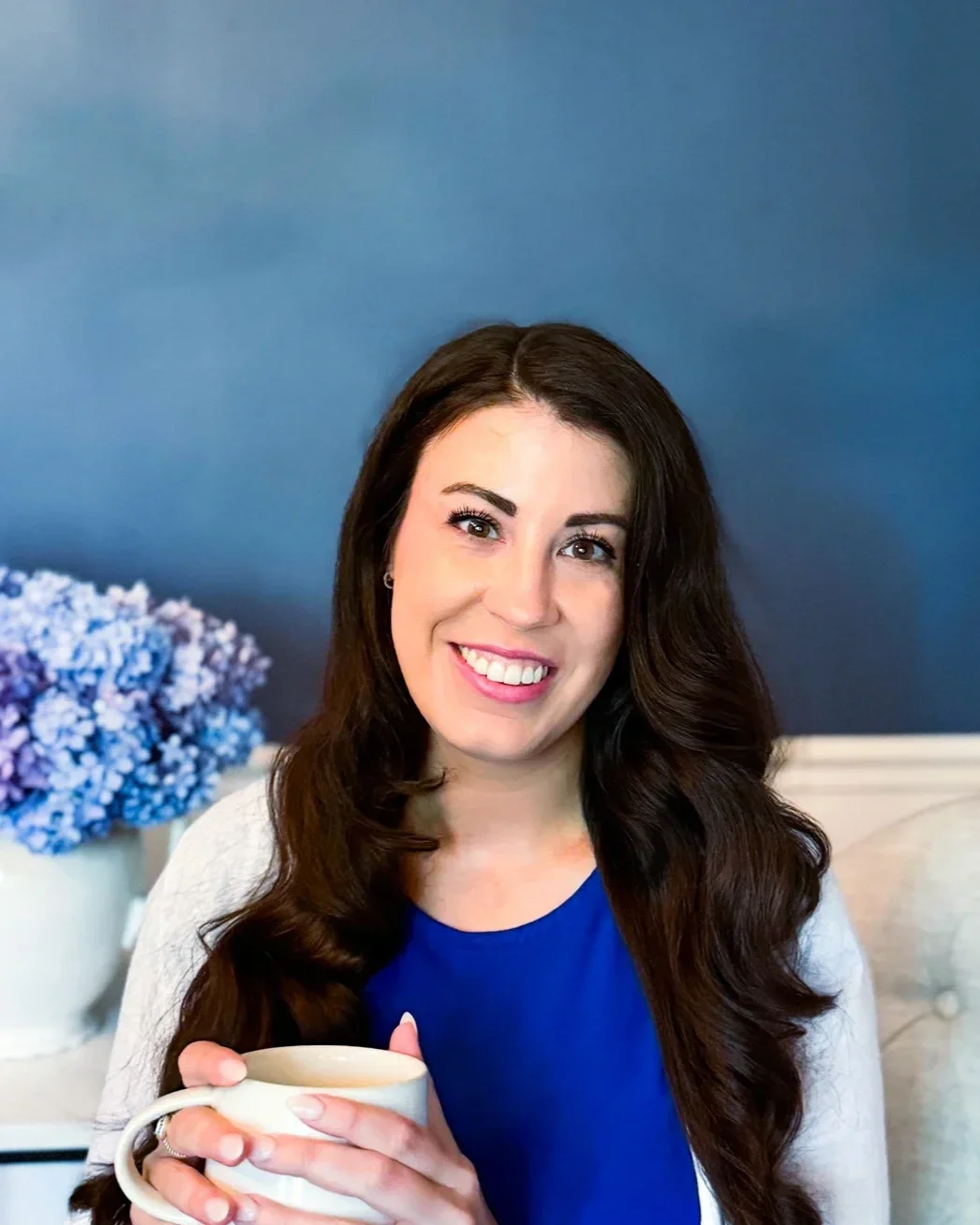 Smiling woman who is an anxiety therapist with long dark hair holding a white mug, standing in front of a blue background and a vase with purple flowers.