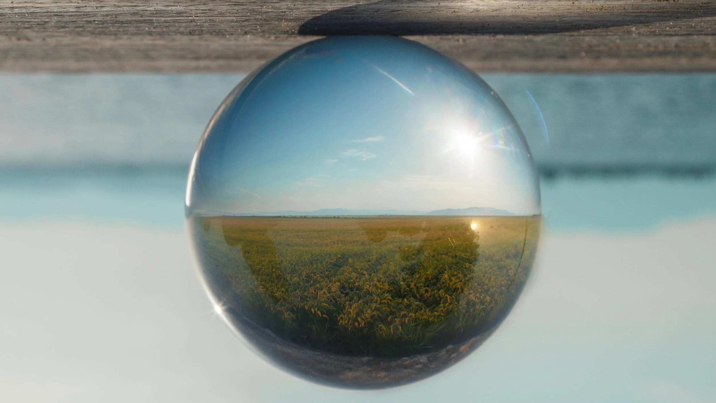 Vista de un campo de cultivo a través de una esfera de cristal, reflejando un cielo despejado con el sol brillante.