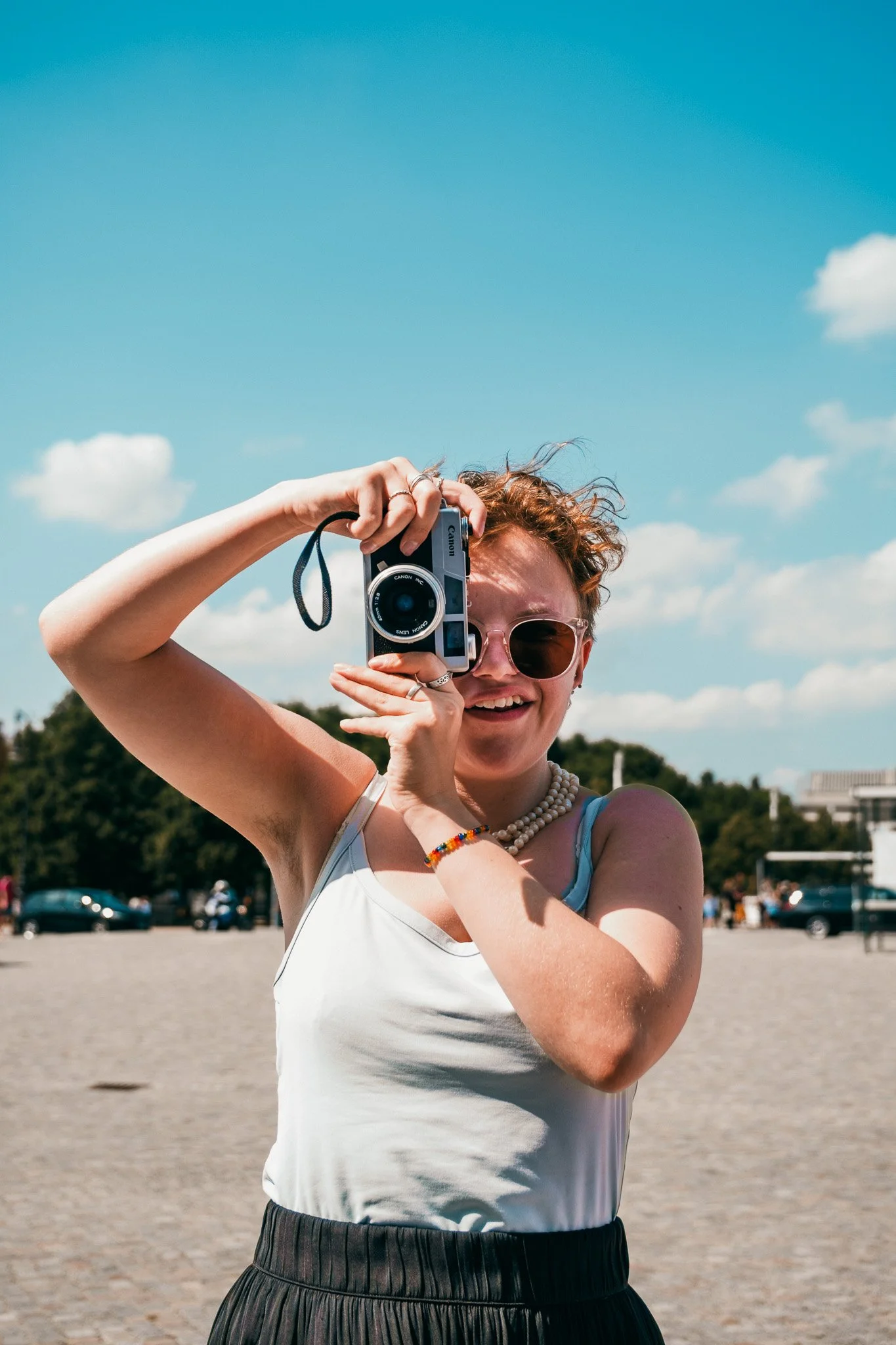 Kvinna med solglasögon, vita kläder och halsband, fotograferar med kamera utomhus mot en klar blå himmel.