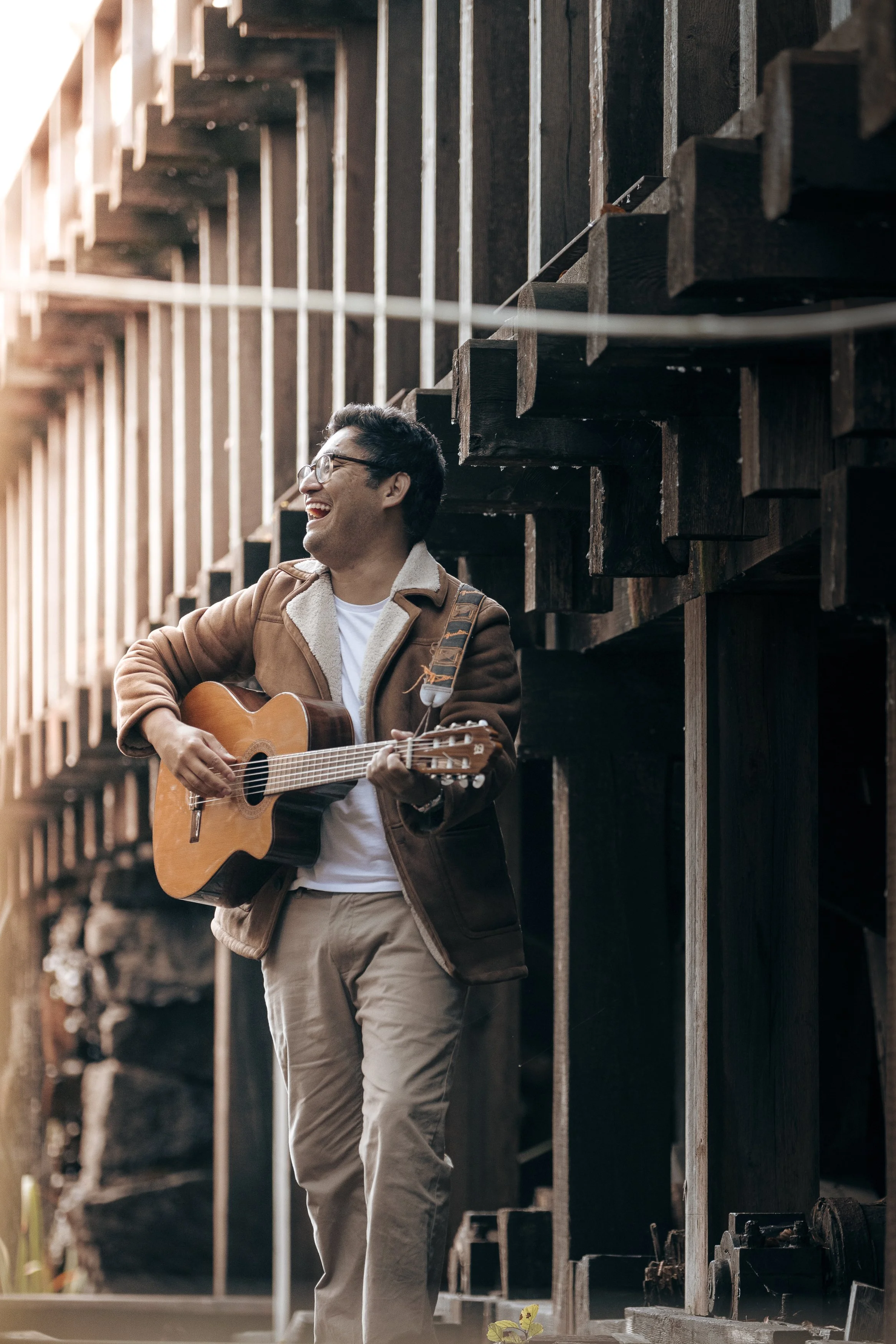 Man som spelar gitarr och skrattar ståendes utomhus bredvid ett trästaket.