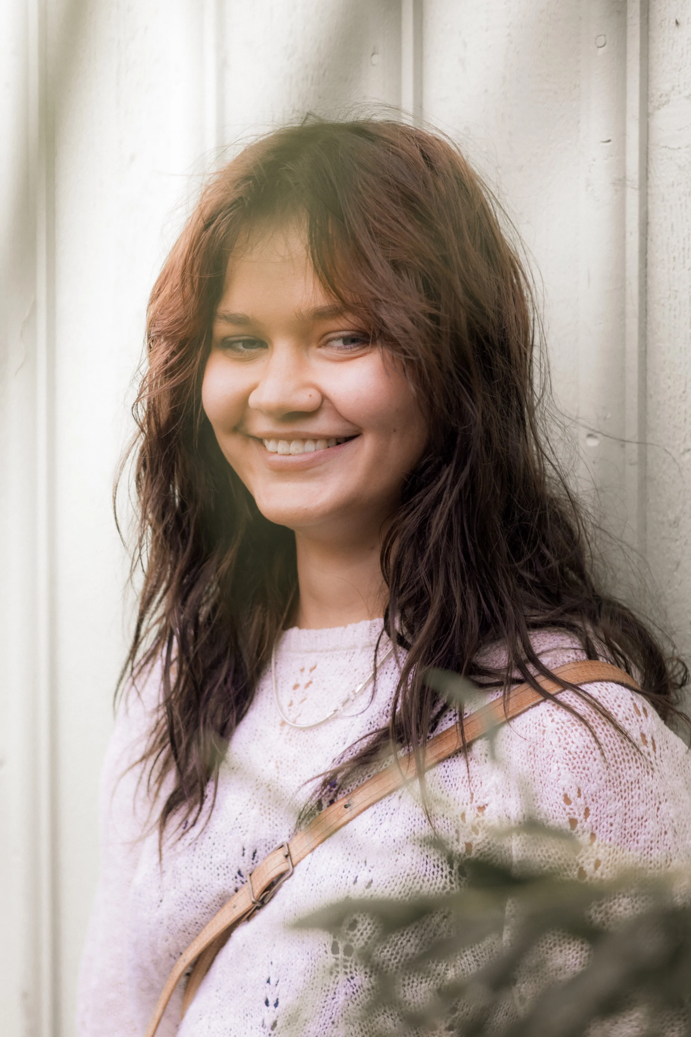 Ung kvinna med mörkt lockigt hår, ler, står vid vägg, bär rosa tröja och skinnväska.