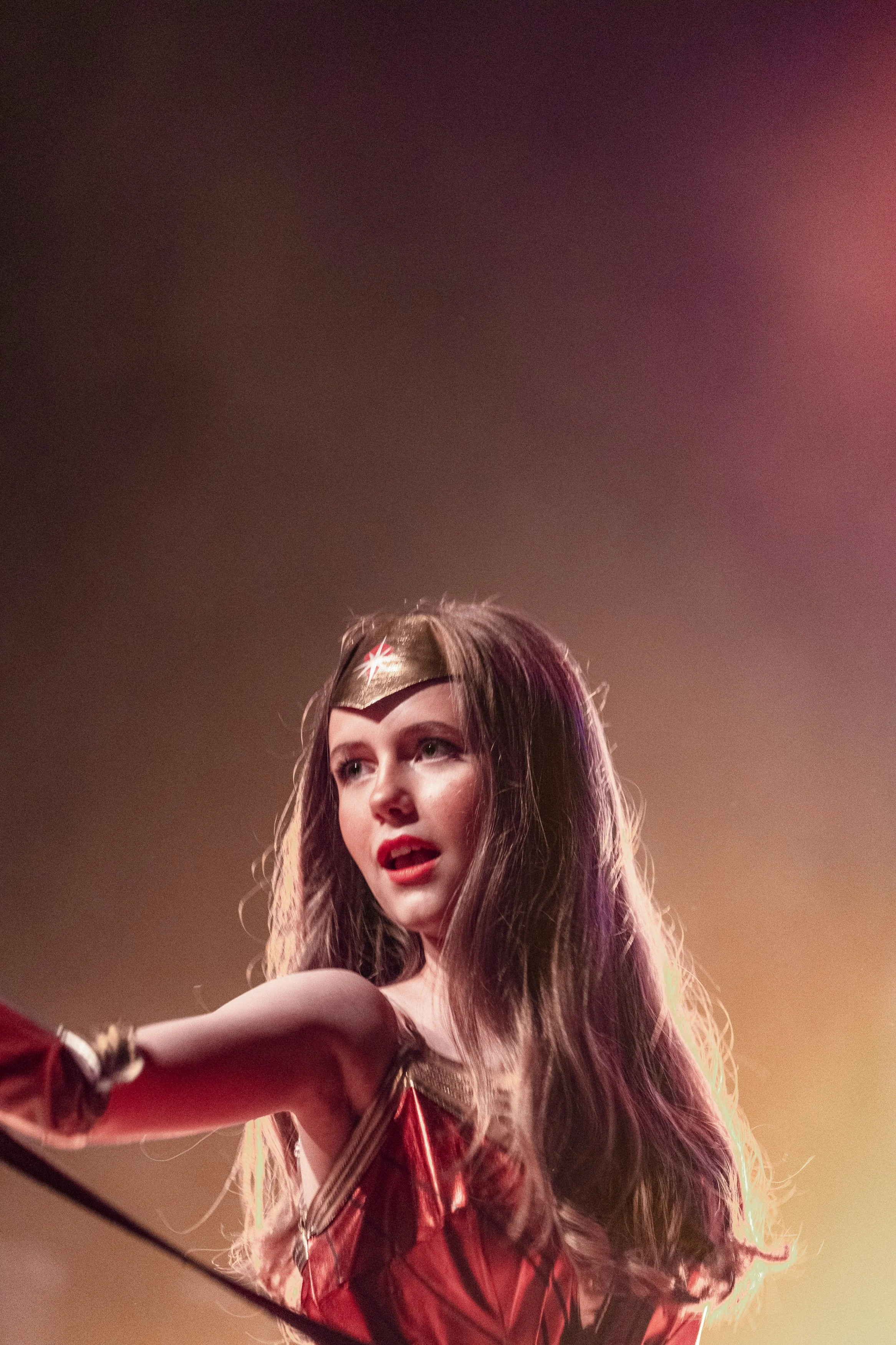 En kvinna i ett Wonder Woman-inspirerat kostym och huvudband står på scen, med ett färgglatt lysande bakgrund.