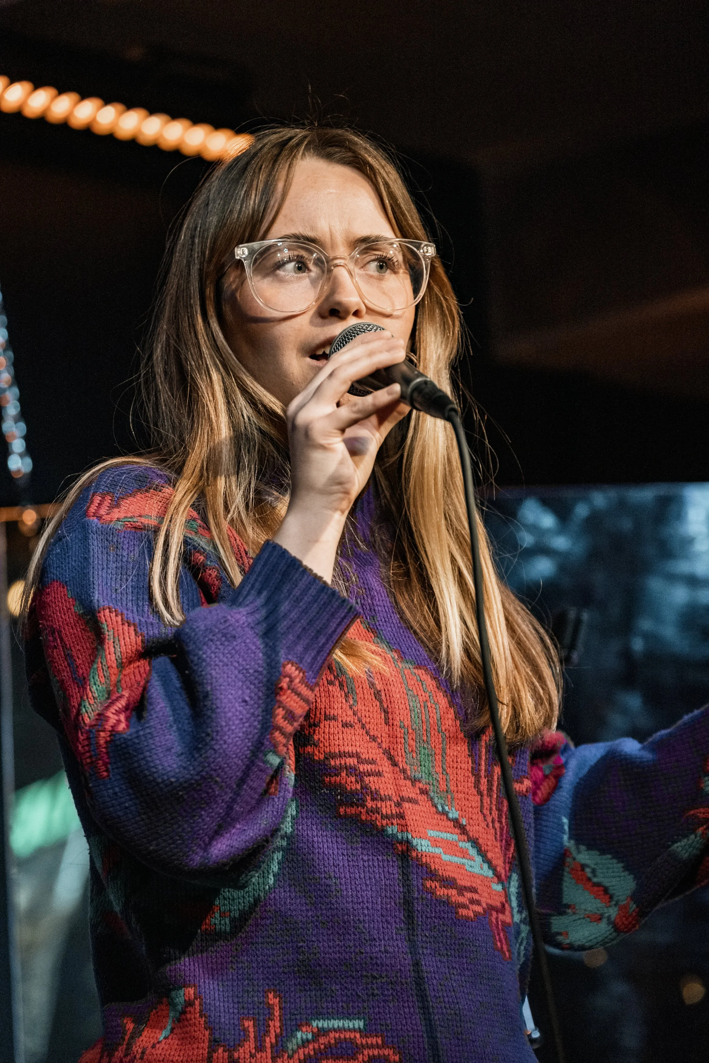 Kvinna med lång brunt hår och stora glasögon, håller i en mikrofon och sjunger på scen.