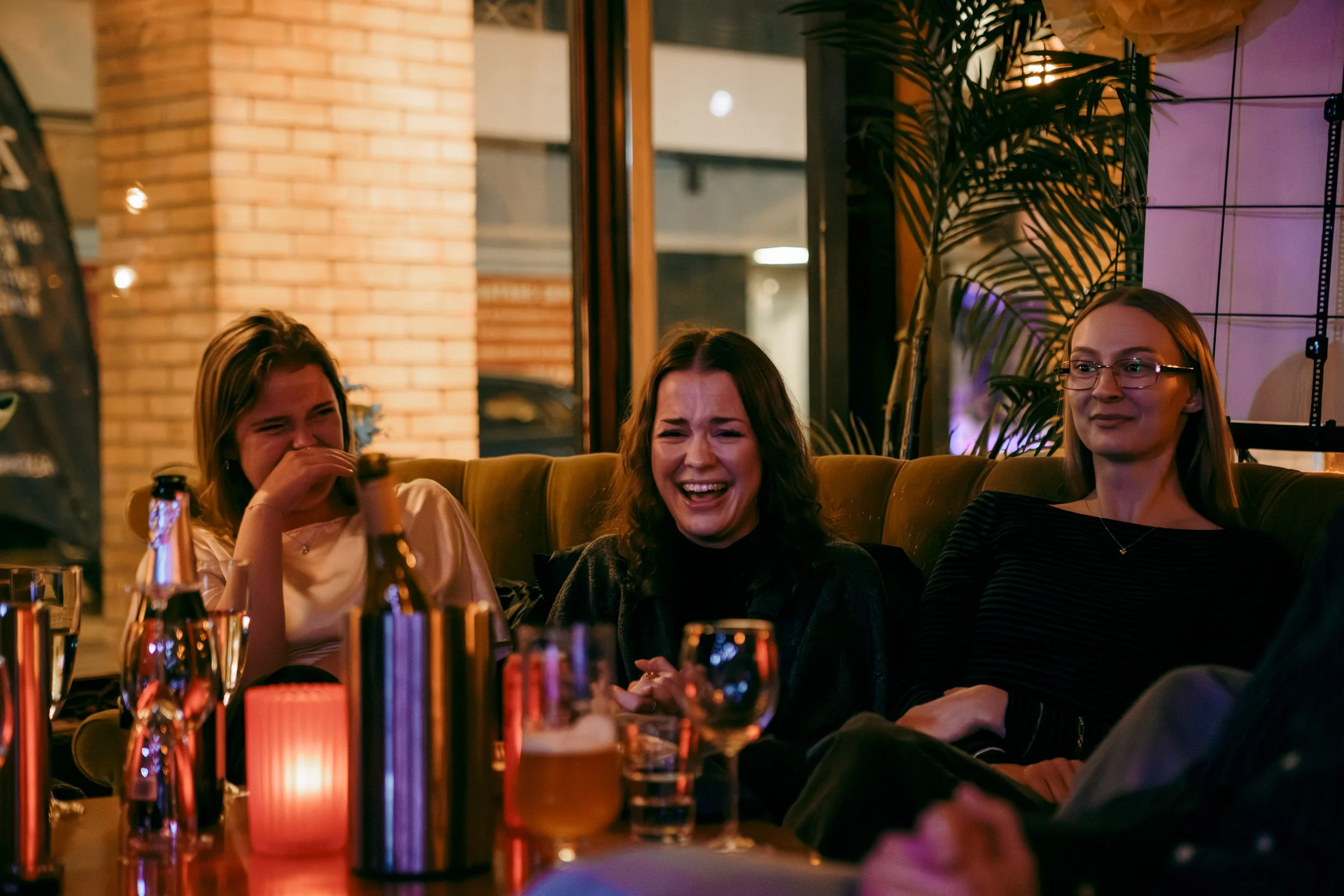 Tre vänner som skrattar och har roligt i en bar eller restaurang, omgiven av drinkar och levande ljus.