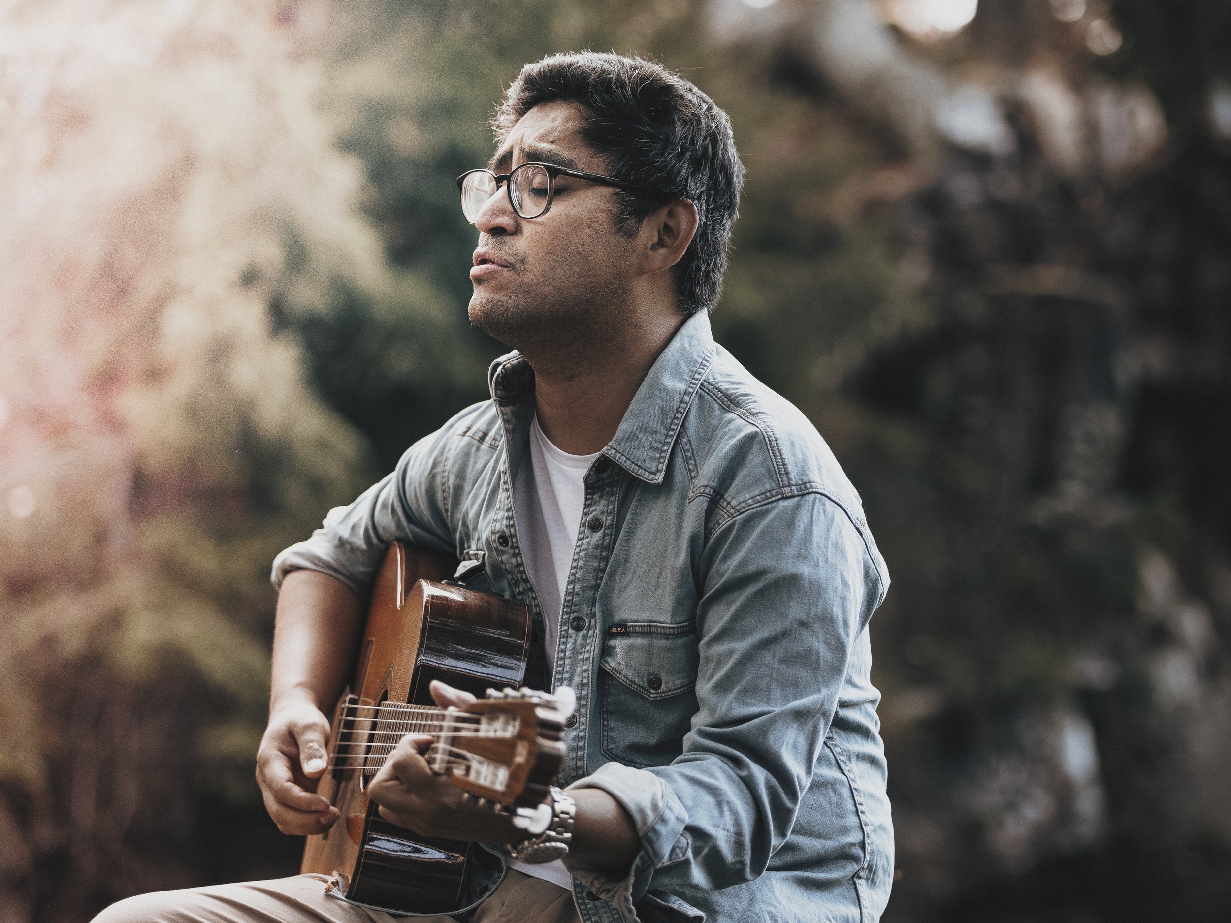 Man med glasögon spelar akustisk gitarr utomhus i en park eller skog.