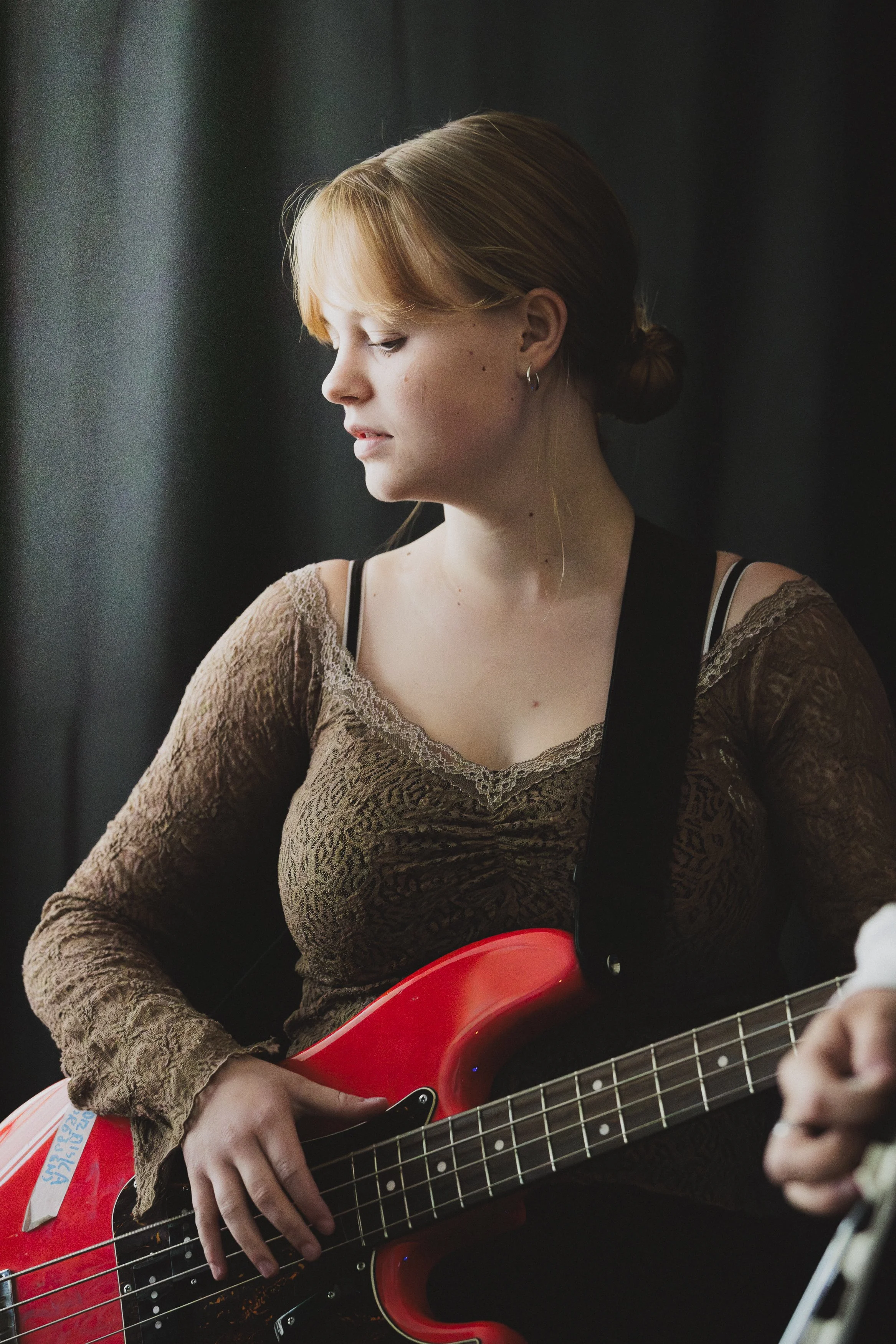 En ung kvinna med rött hår som spelar elbas, hon bär en brunt ljust tröja med spetsdetaljer och har ett svart band över ena axeln.